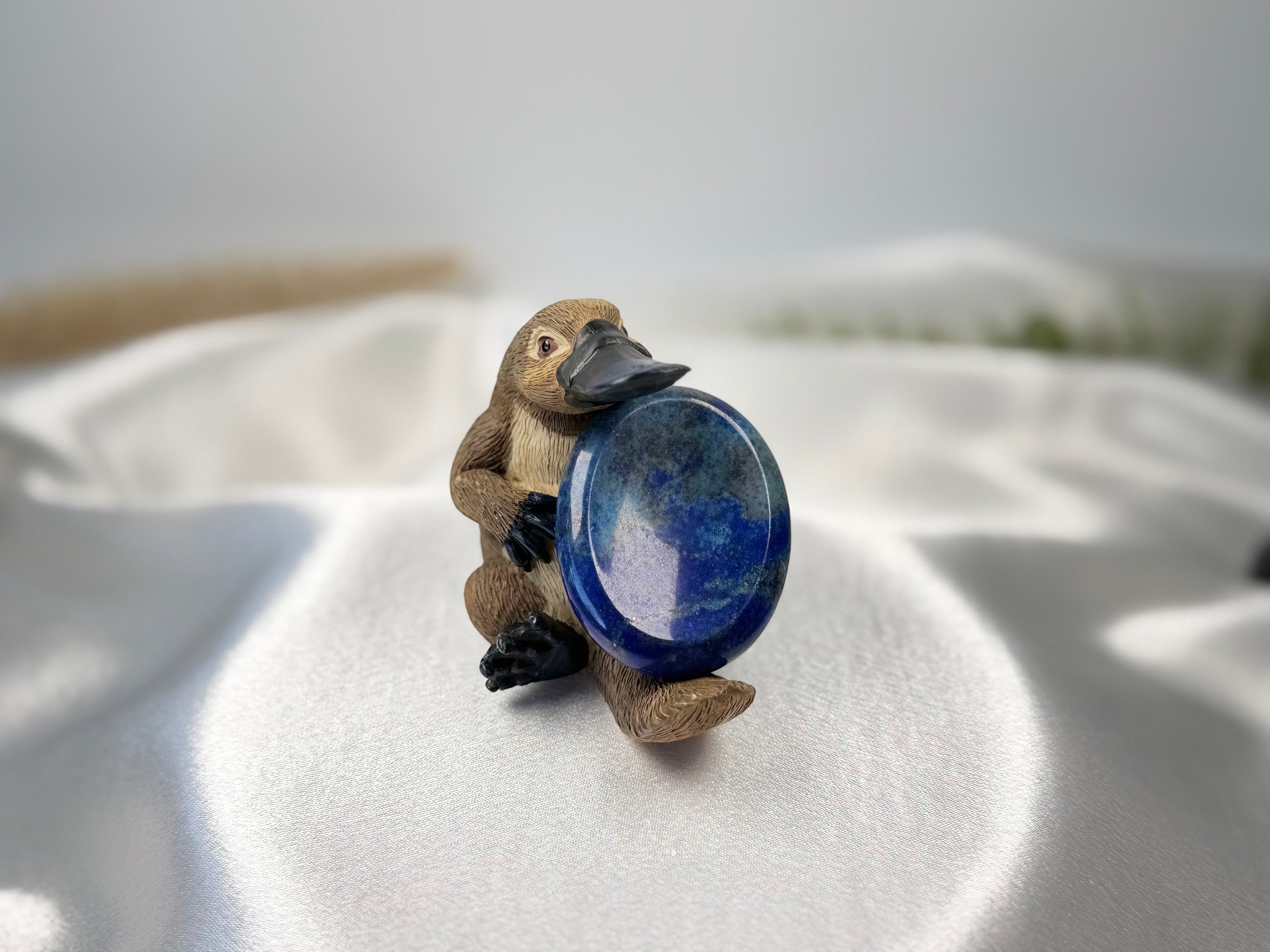 Treasured Energies Lapis Lazuli worry stone held by a small platypus figurine on a white satin fabric background.