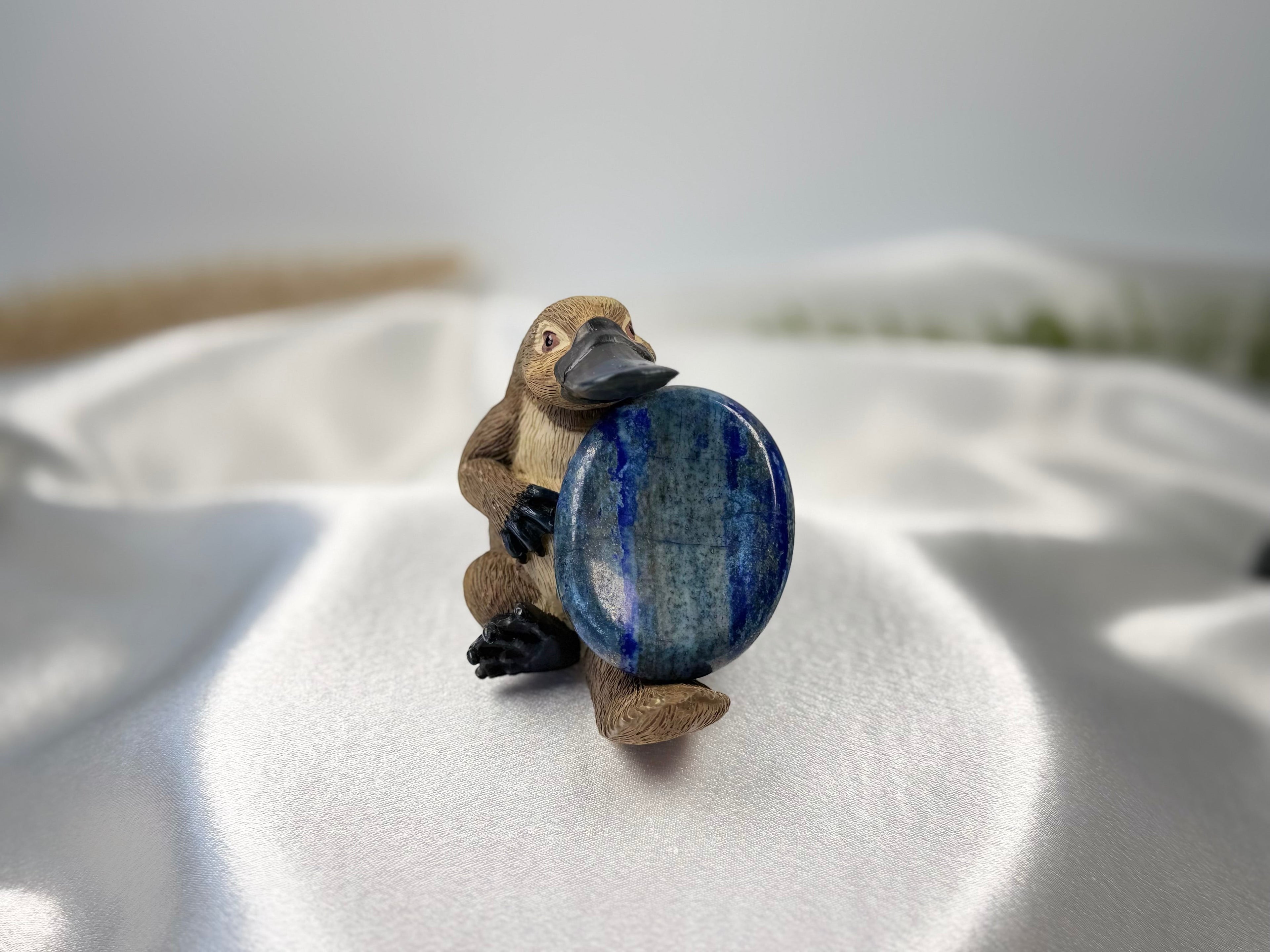 Treasured Energies deep blue Lapis Lazuli worry stone held by a small platypus figurine on a white satin background