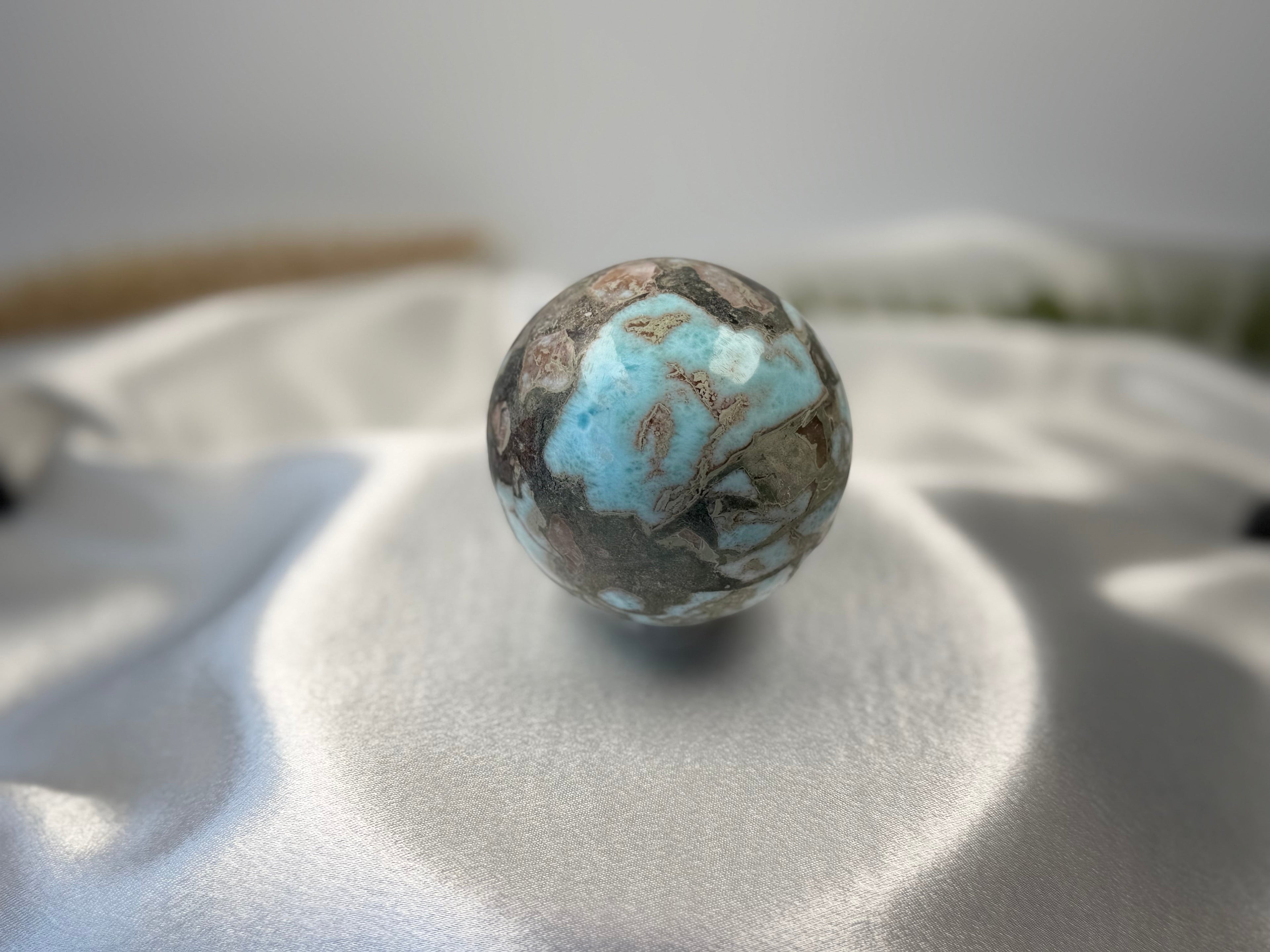 Treasured Energies polished Larimar crystal sphere with blue and brown patterns on a soft white satin fabric background.