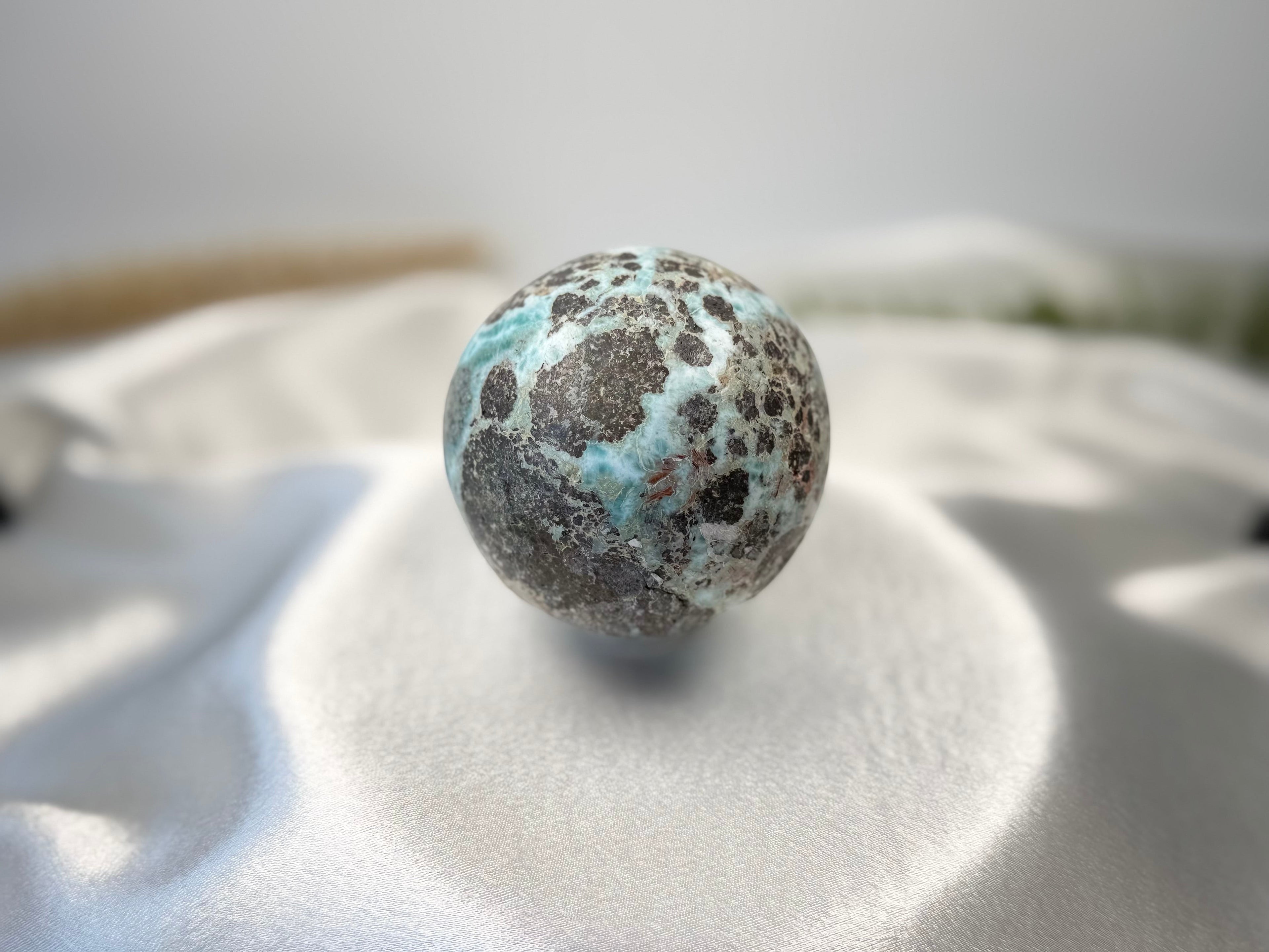Treasured Energies polished Larimar sphere with blue and brown patterns resting on a soft white satin fabric.