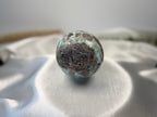 Treasured Energies Larimar crystal sphere with blue and dark brown patterns resting on a white satin fabric background.