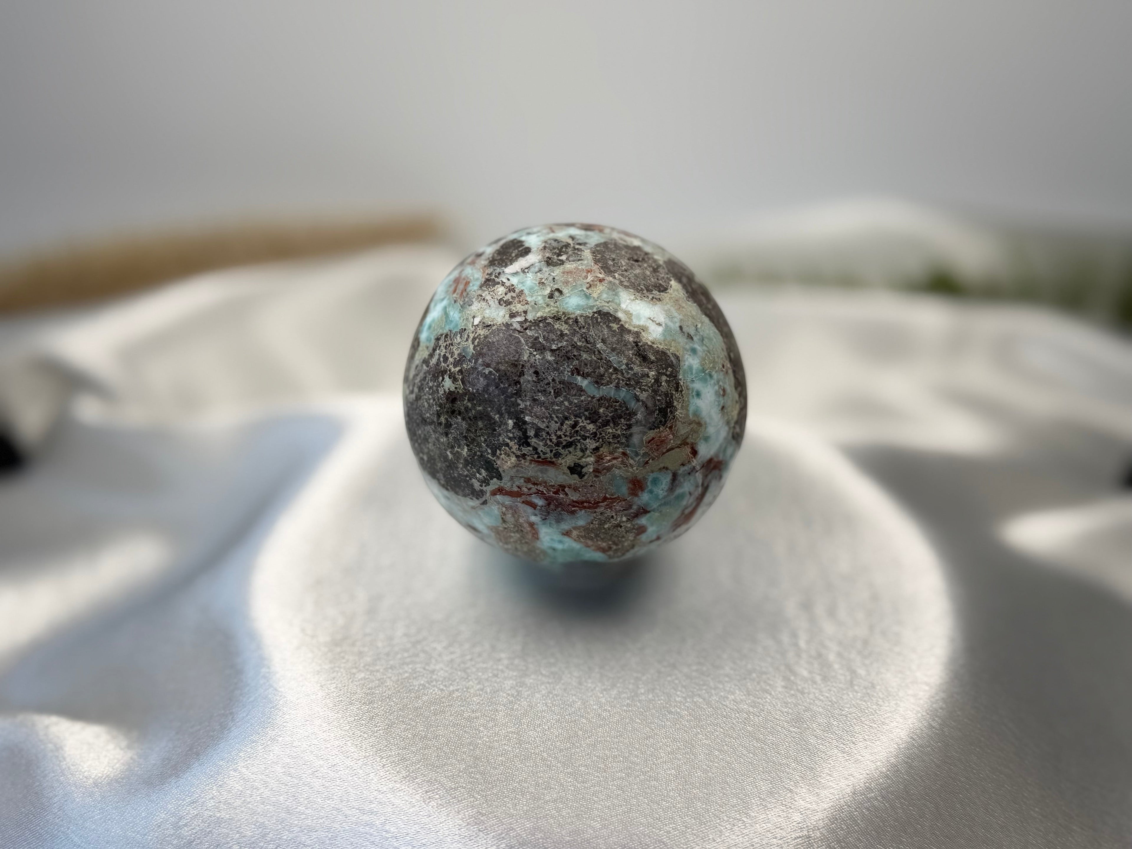 Treasured Energies Larimar crystal sphere with blue and dark brown patterns resting on a white satin fabric background.