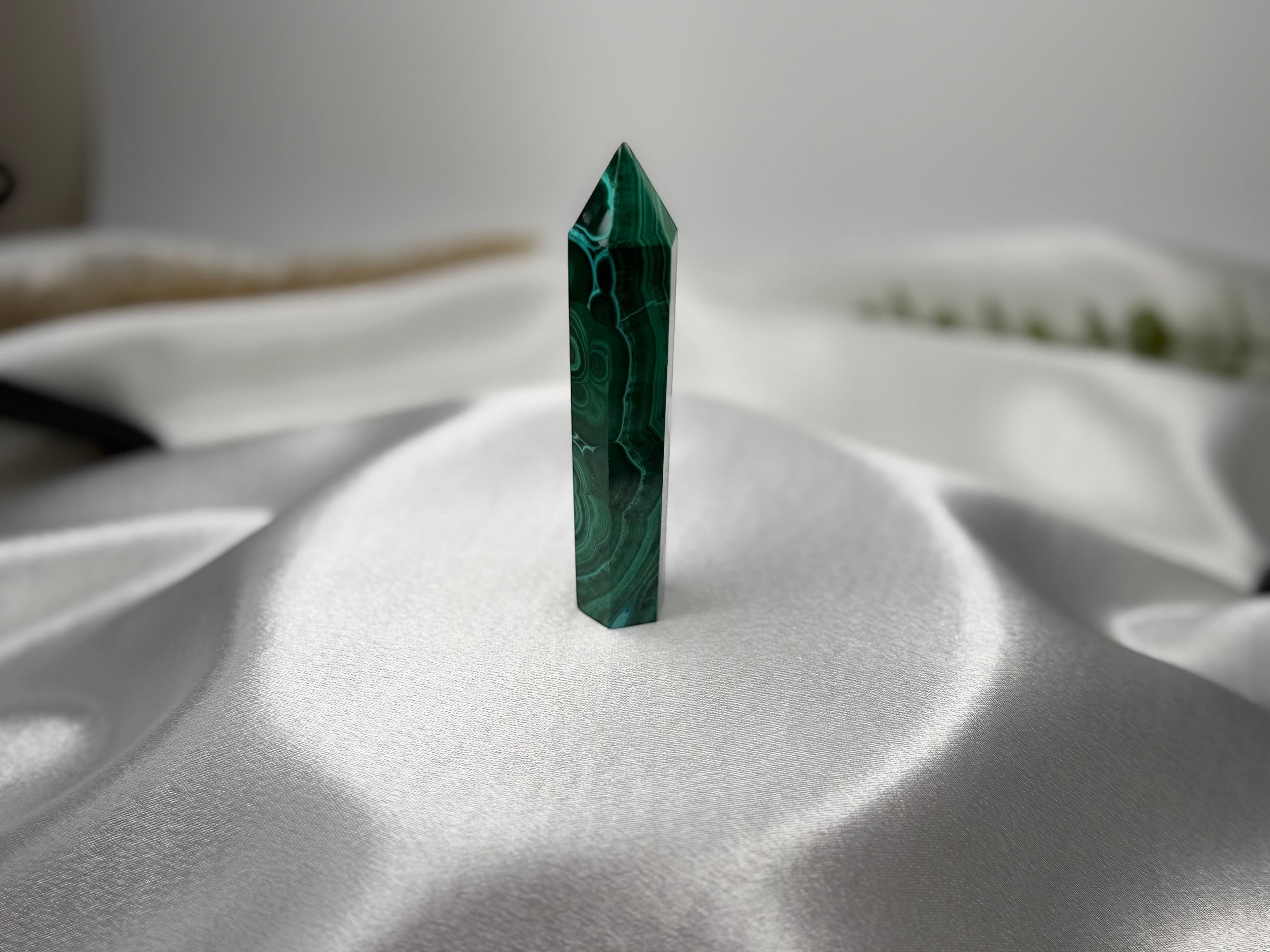 Treasured Energies polished malachite crystal tower with deep green banding on a soft white satin background.