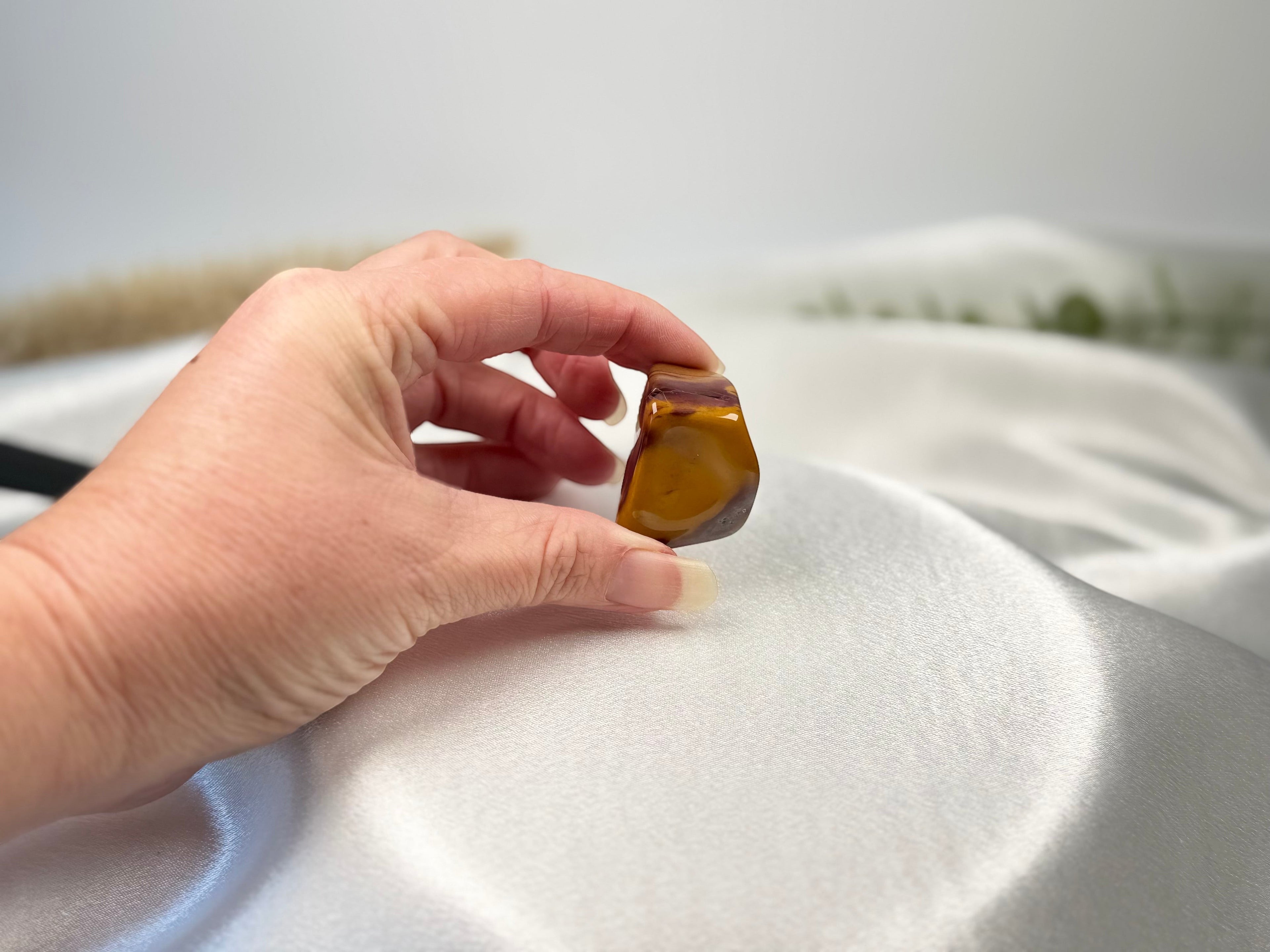 Treasured Energies large polished Mookaite tumbled stone with earthy red, burgundy, and yellow tones held in a hand.