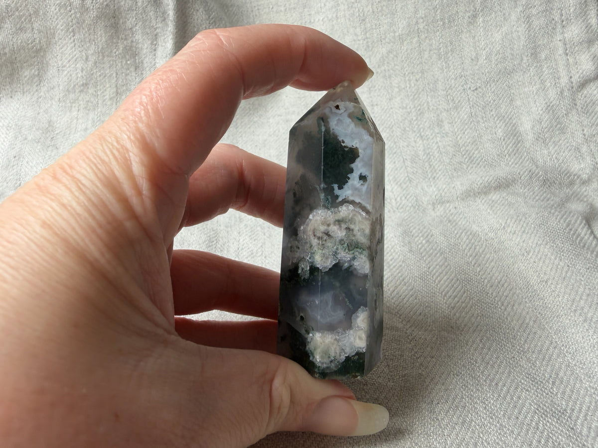 Treasured Energies polished Moss Agate crystal tower held in hand, showing green moss-like inclusions and grey quartz.