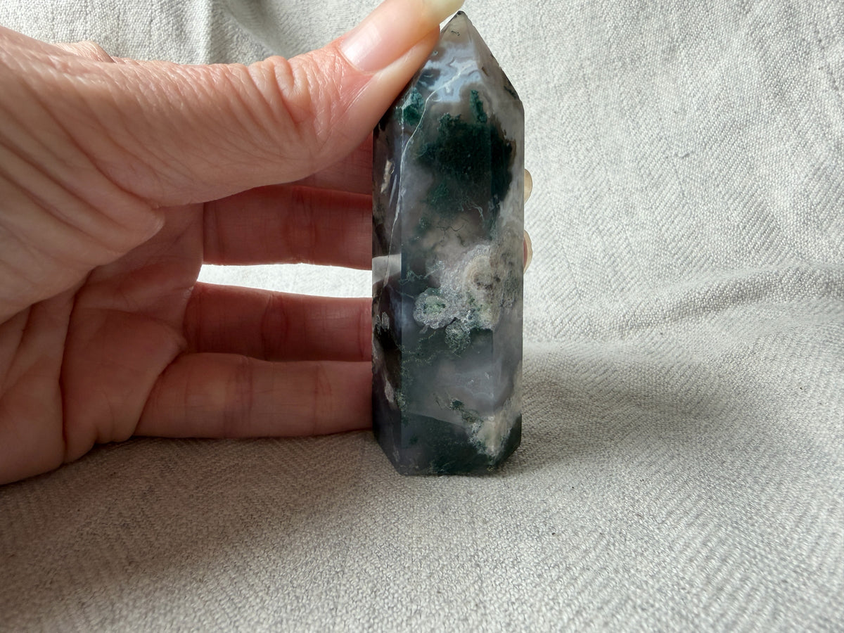 Treasured Energies polished Moss Agate crystal tower with green moss-like inclusions held against a linen background.