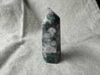 Treasured Energies polished Moss Agate crystal tower point with green moss-like inclusions on a textured linen fabric.
