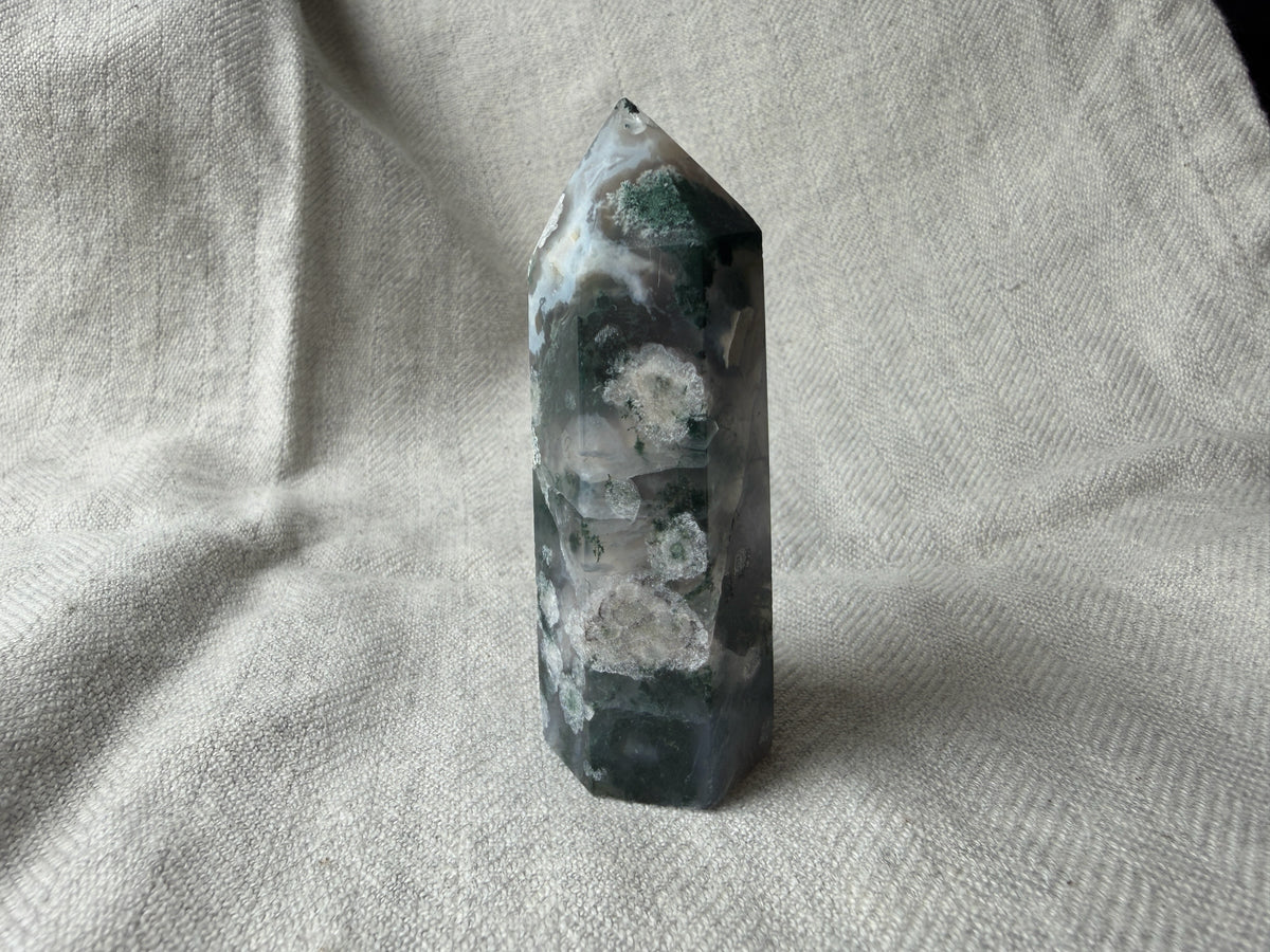 Treasured Energies polished Moss Agate crystal tower point with green moss-like inclusions on a textured linen fabric.