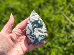 Treasured Energies polished moss agate flame crystal sculpture held in hand with green garden background
