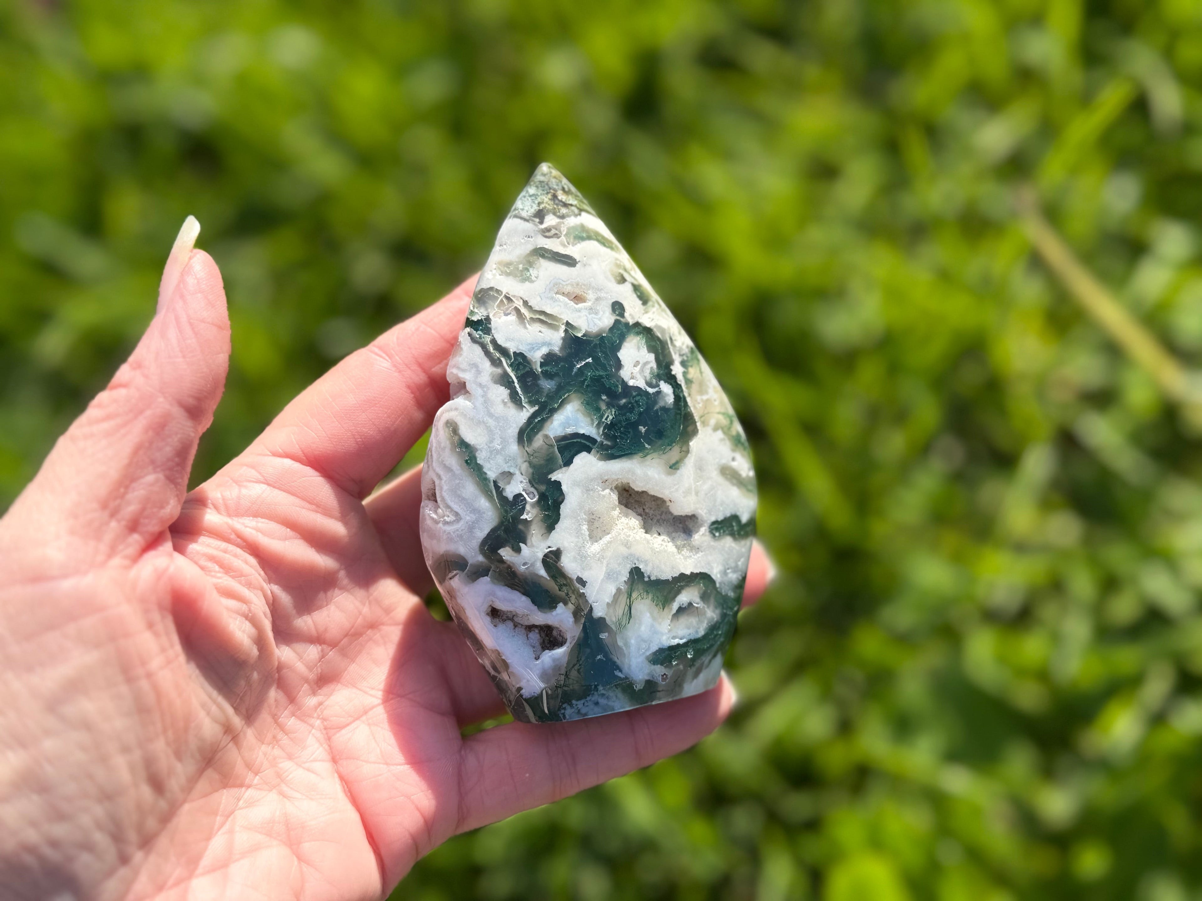 Treasured Energies polished moss agate flame crystal sculpture held in hand with green garden background