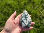 Treasured Energies polished moss agate flame crystal sculpture held in hand against a blurred green garden background