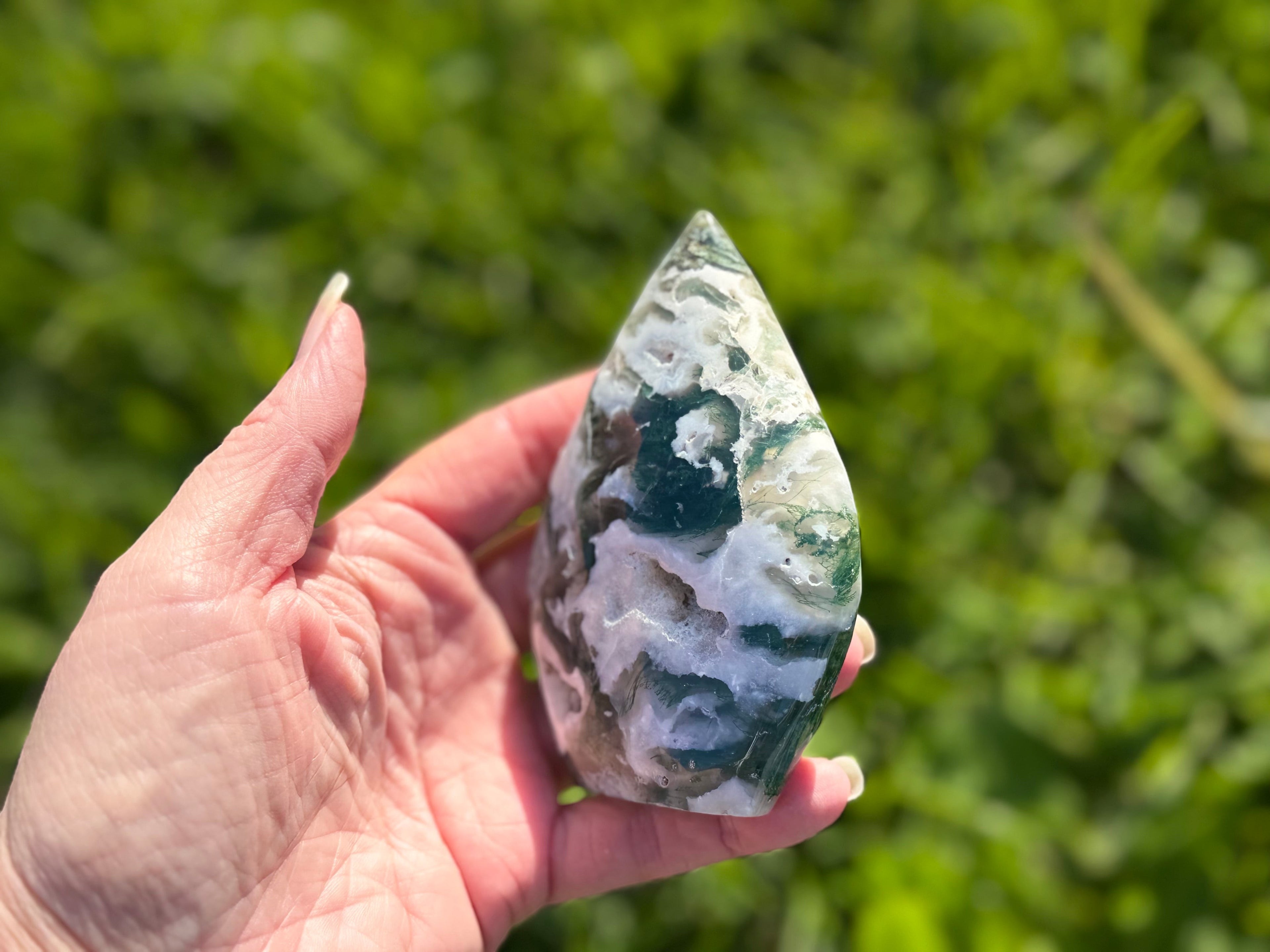Treasured Energies polished moss agate flame crystal sculpture held in a hand against a blurred green garden background