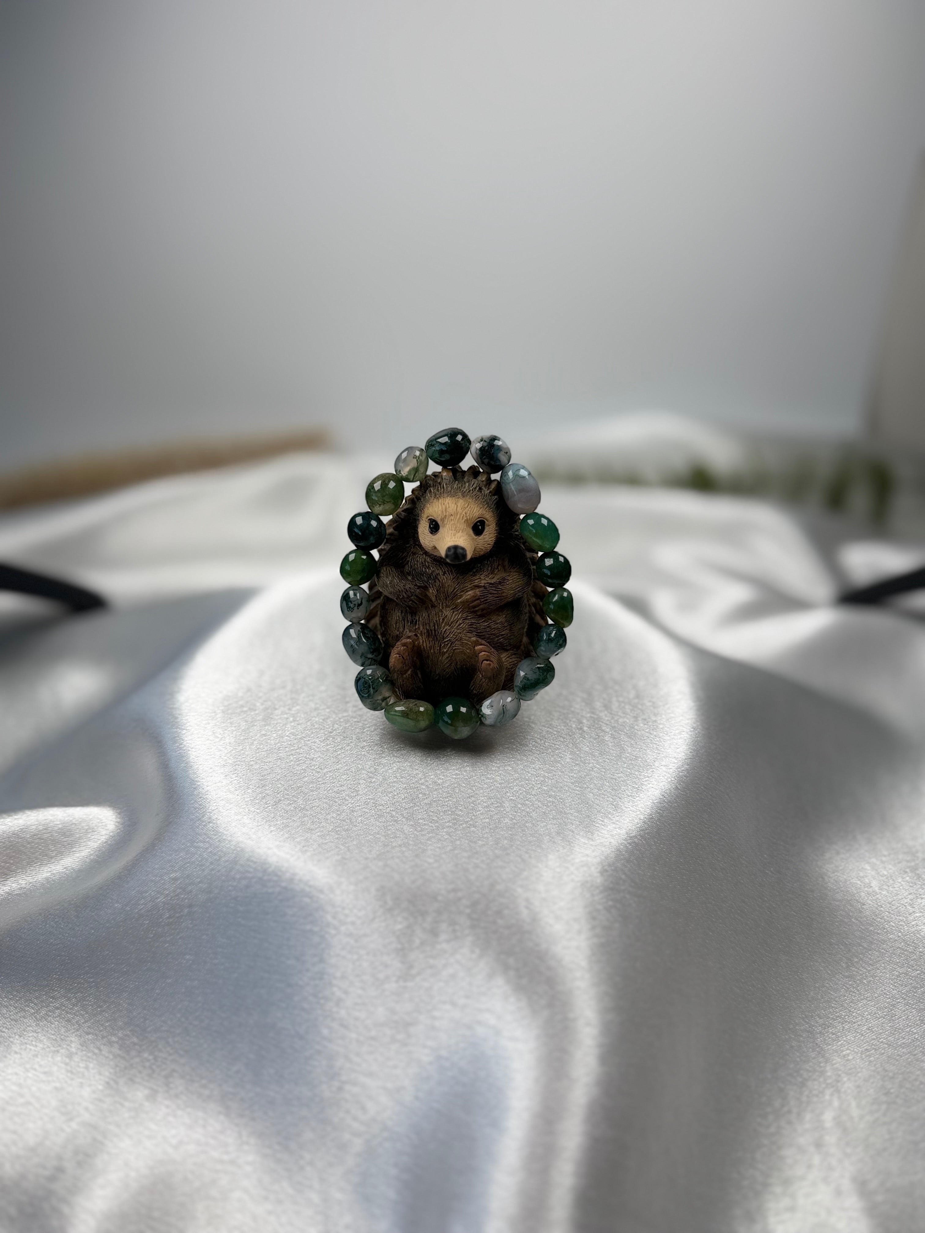 Treasured Energies moss agate crystal bead bracelet encircling a small brown hedgehog figurine on white satin fabric