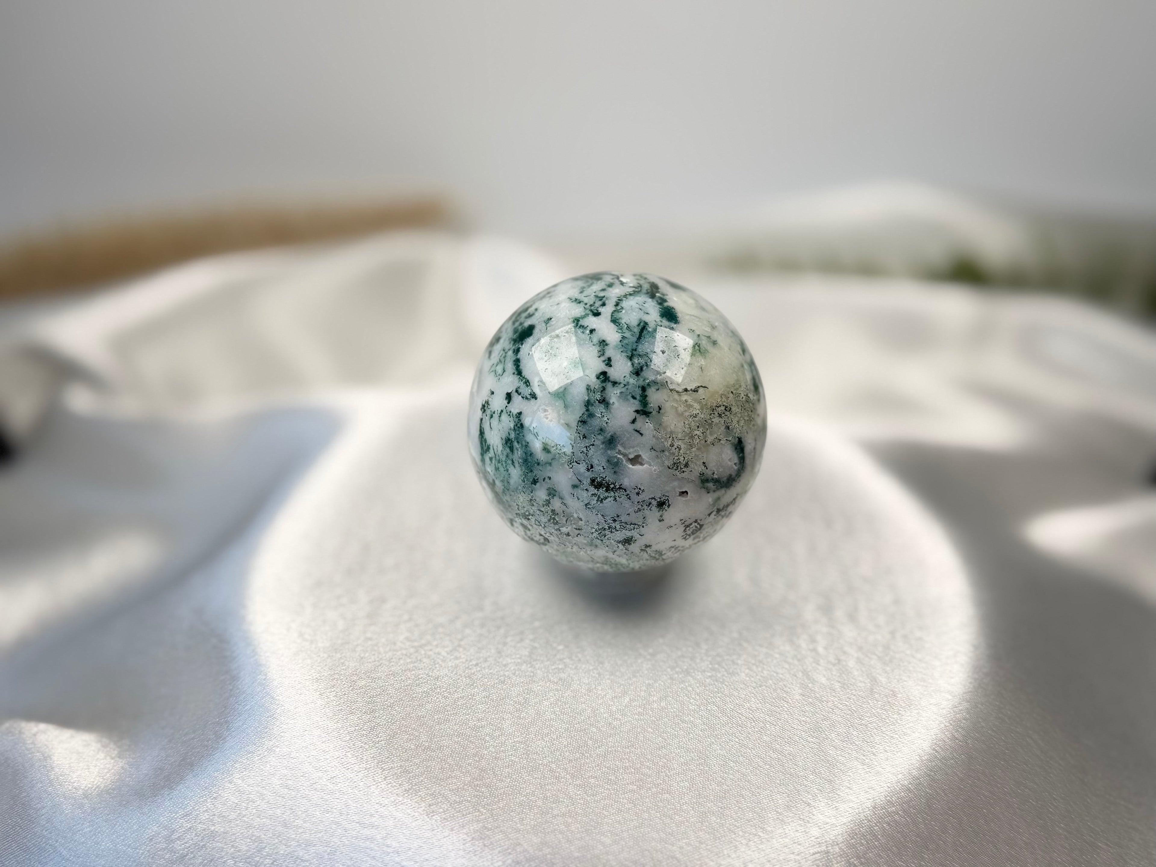 Treasured Energies polished moss agate crystal sphere with green moss-like inclusions on a white satin background.