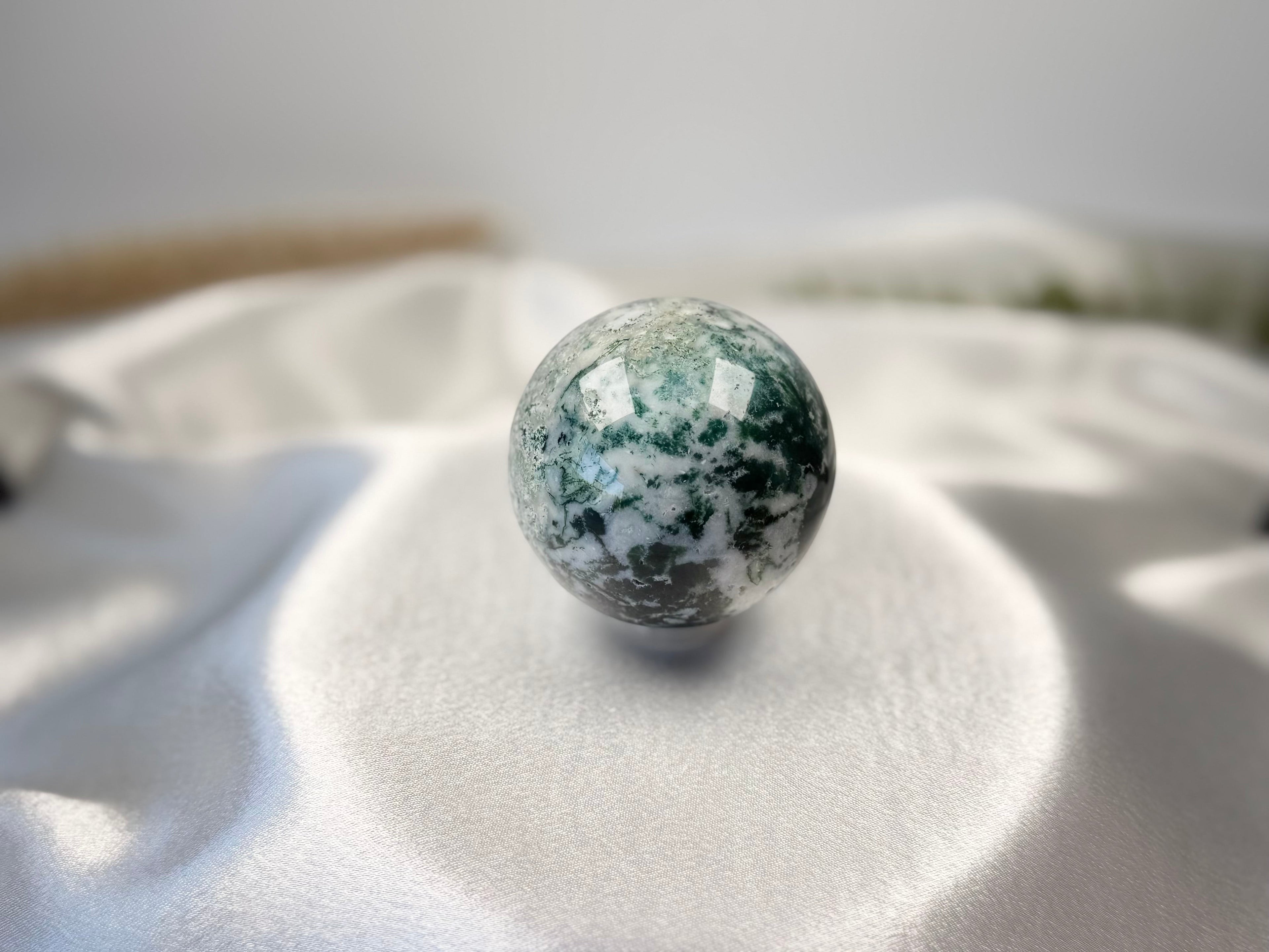 Treasured Energies polished moss agate crystal sphere with green and white earthy inclusions on a white satin cloth.