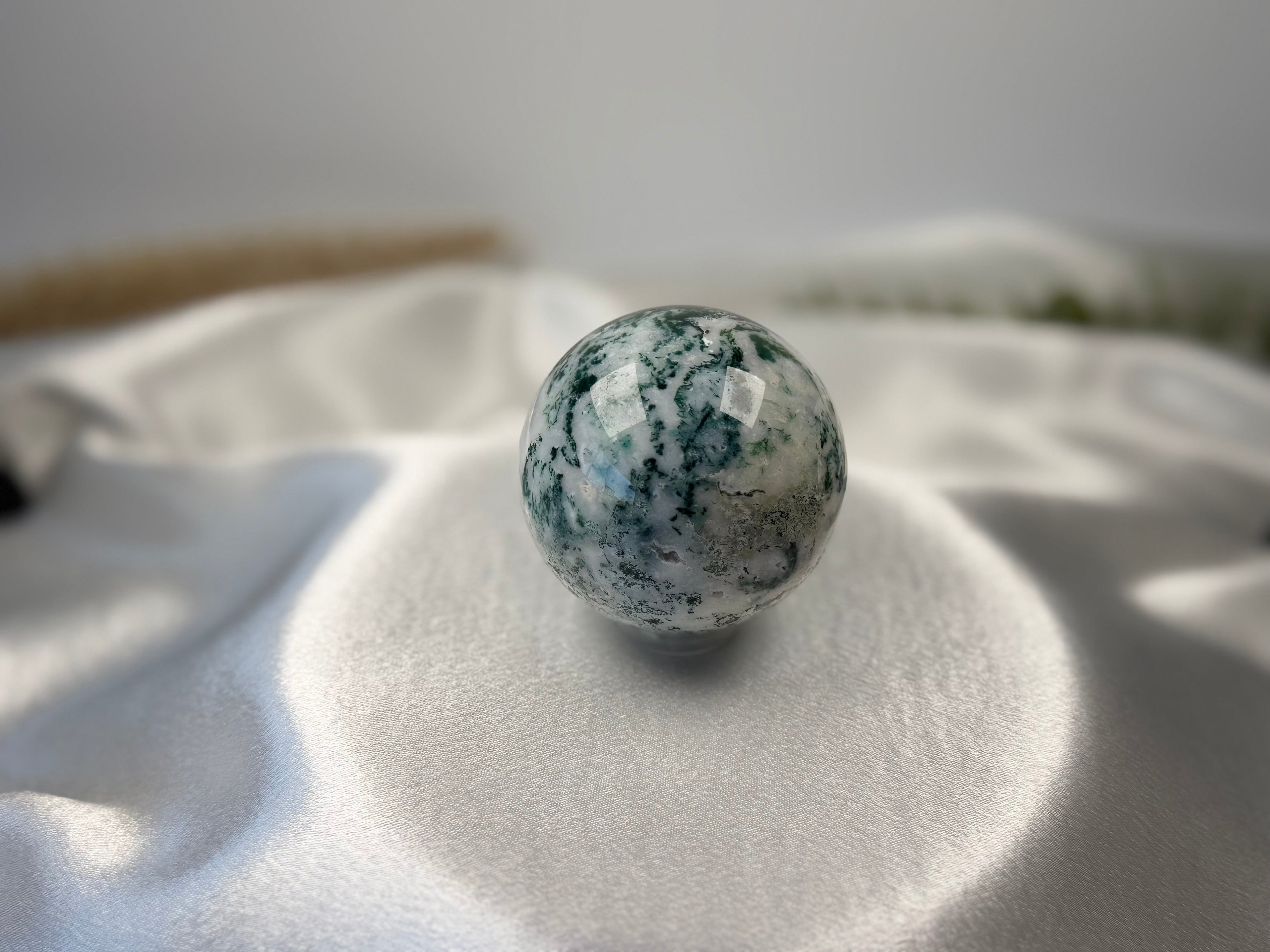 Treasured Energies polished moss agate crystal sphere with green earthy inclusions on a white satin background.