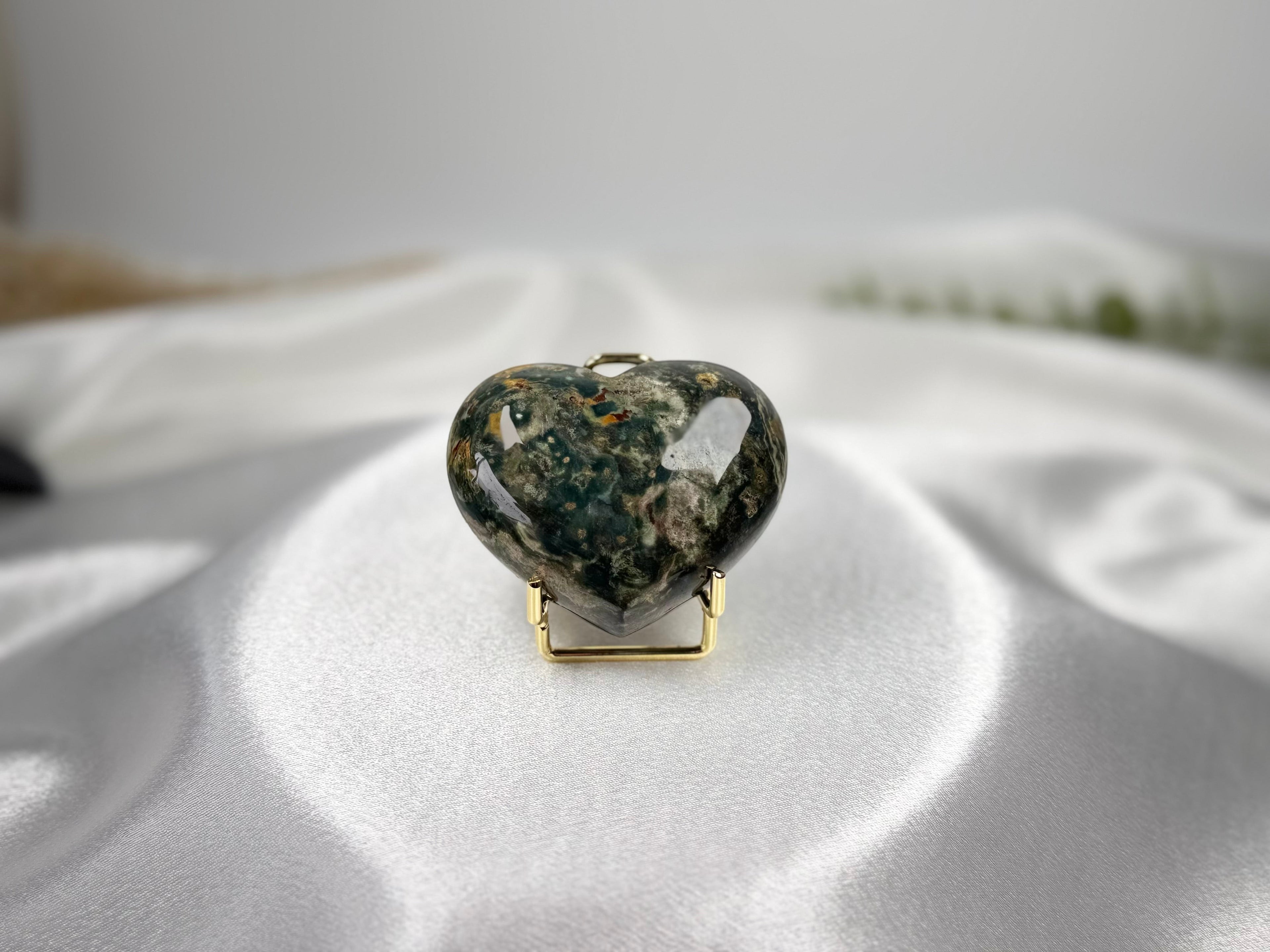 Treasured Energies large polished Ocean Jasper crystal heart on a gold stand against a white satin background.