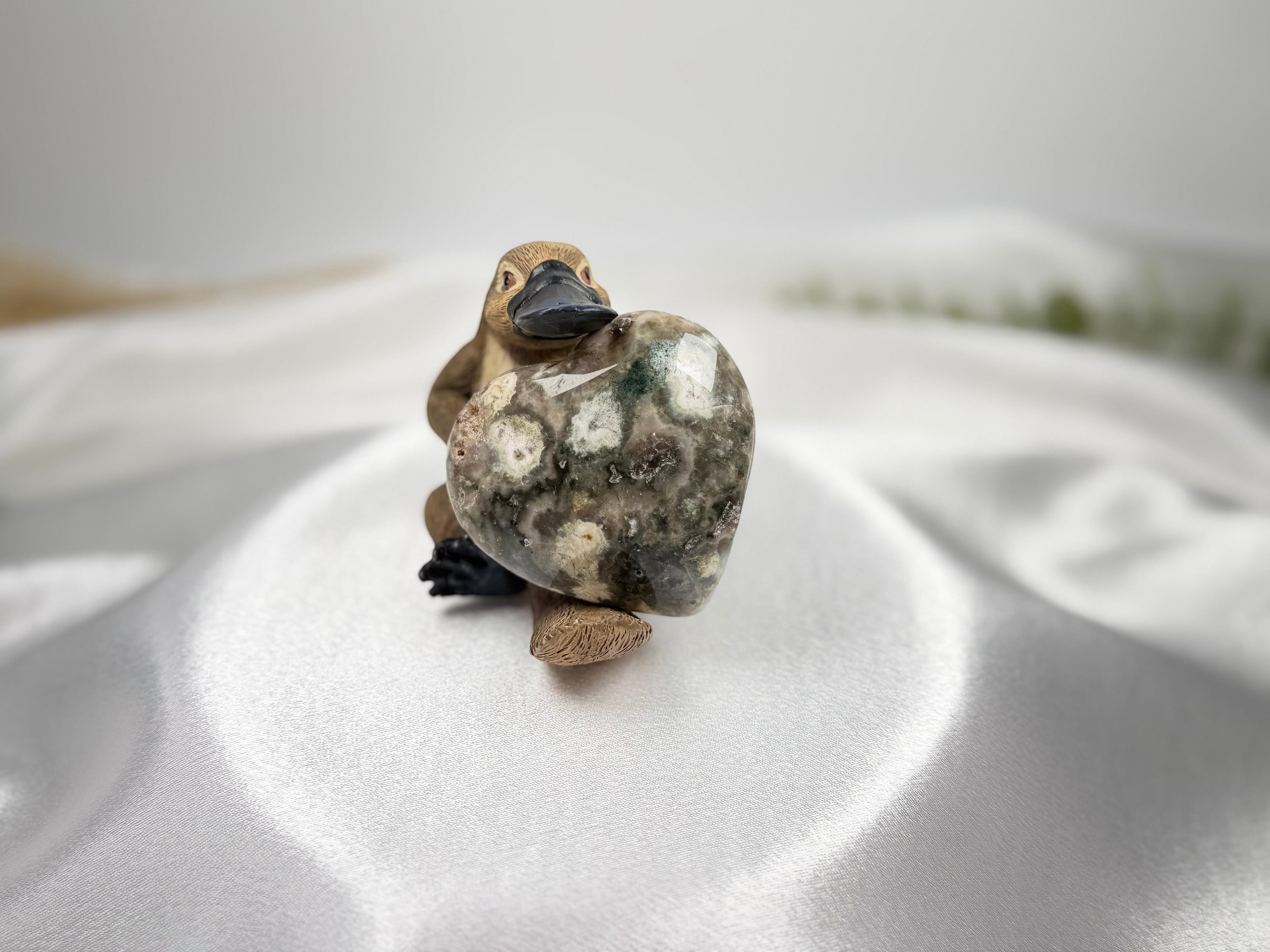 Treasured Energies large polished Ocean Jasper crystal heart held by a small platypus figurine on white satin fabric.