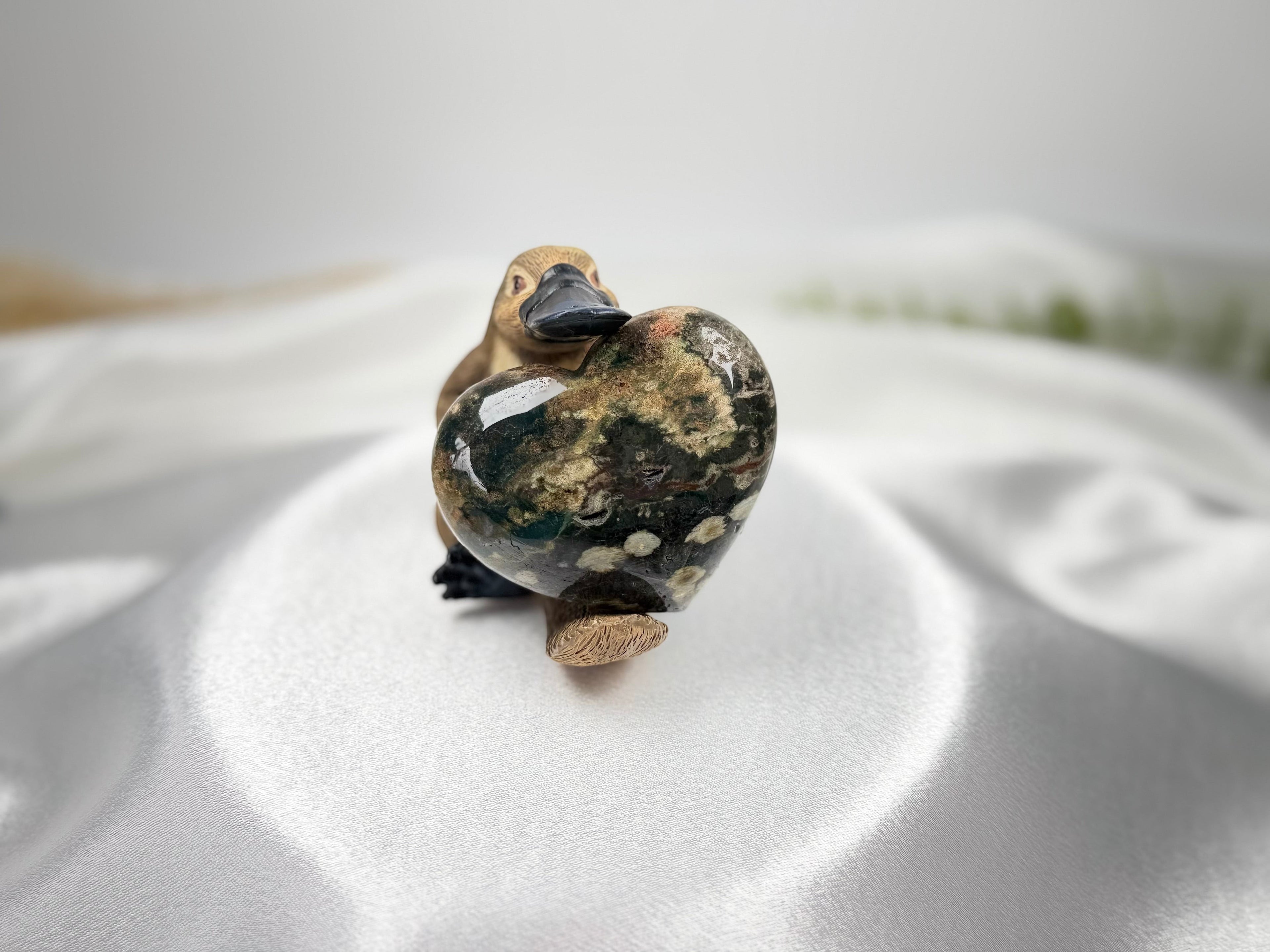 Treasured Energies large polished Ocean Jasper crystal heart held by a small wooden platypus figurine on white silk.