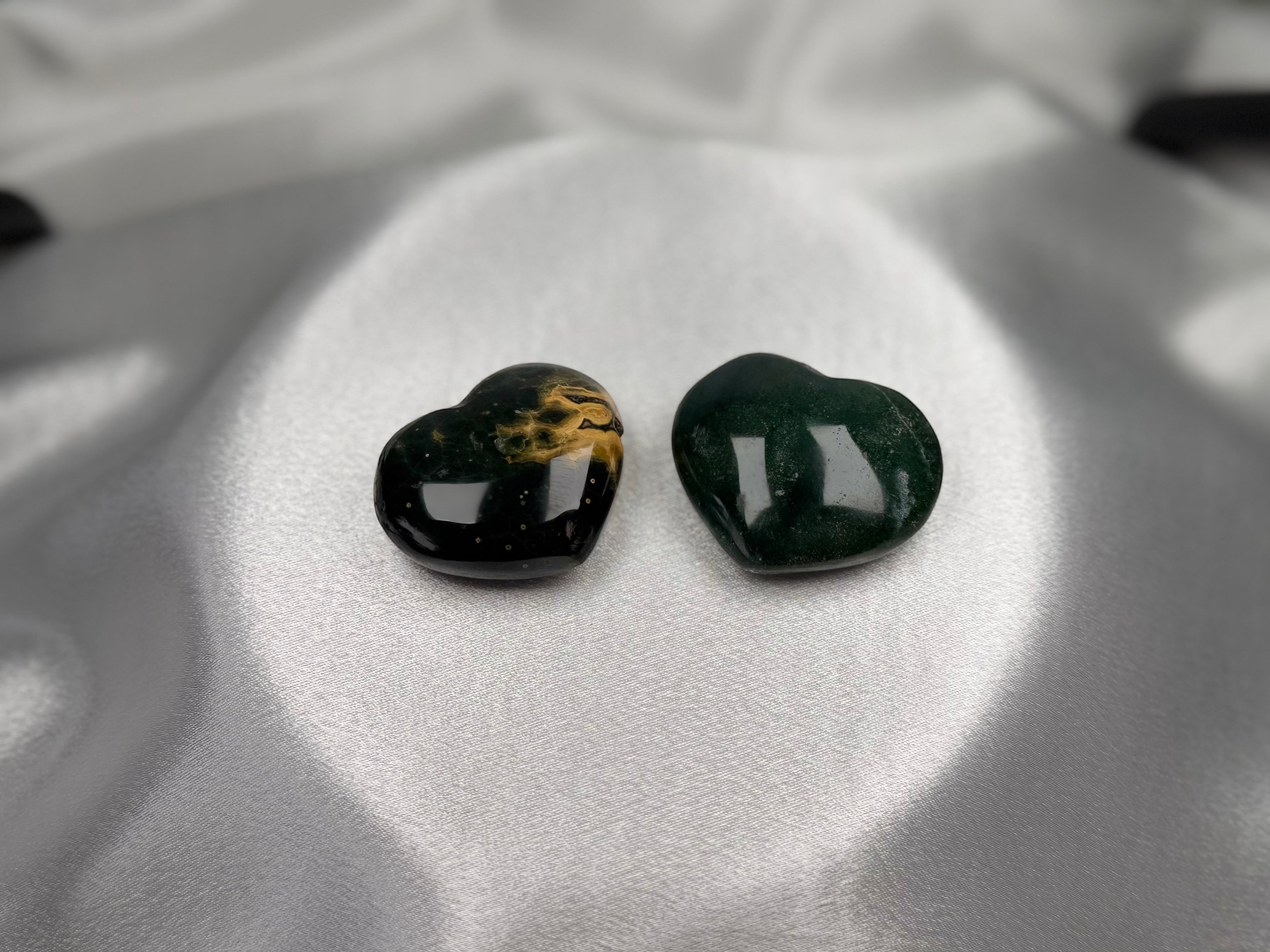 Two Treasured Energies polished Ocean Jasper heart crystals with dark green and gold patterns on white satin fabric.