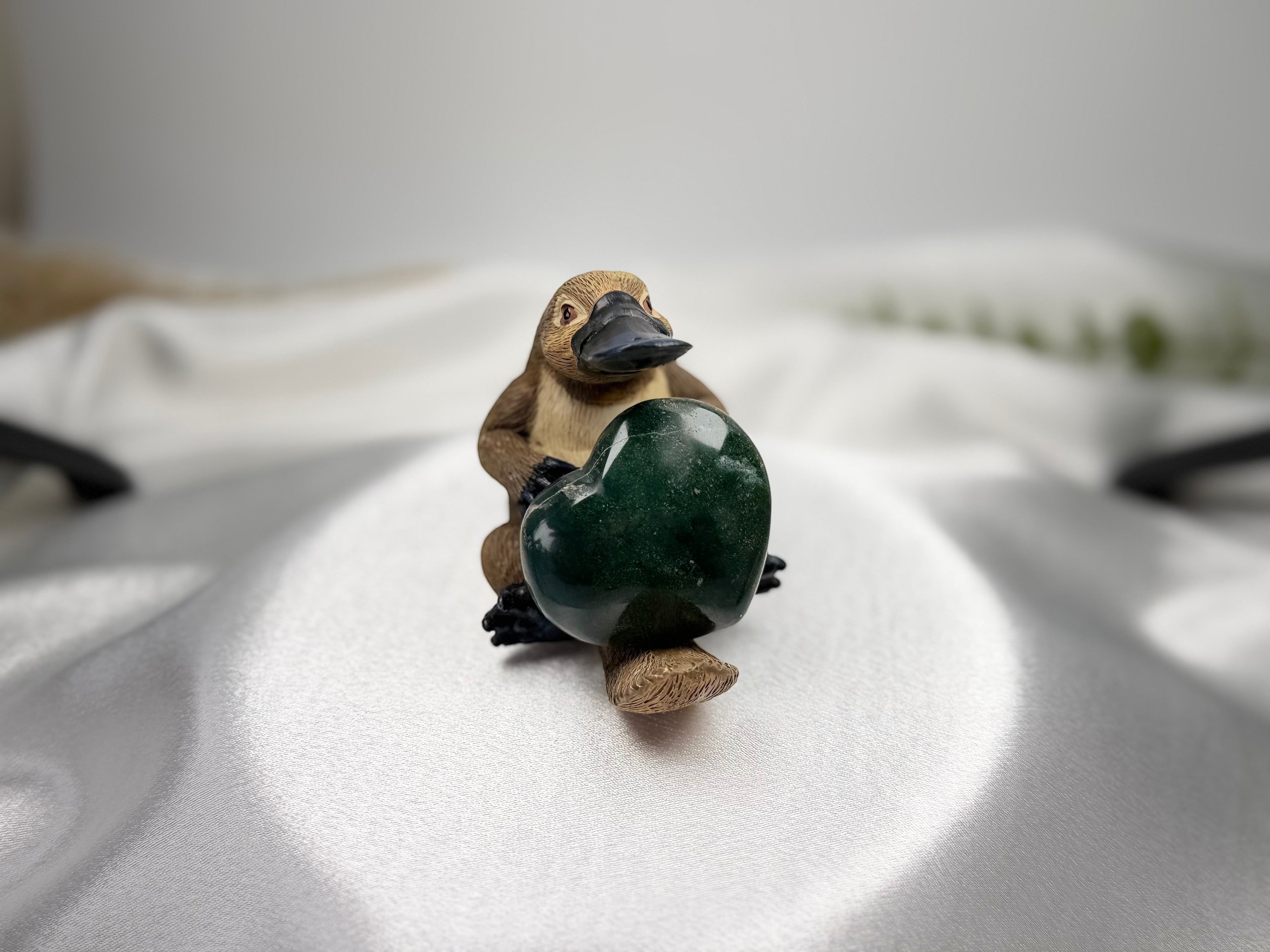 Treasured Energies small polished green Ocean Jasper heart held by a cute carved platypus figurine on white fabric