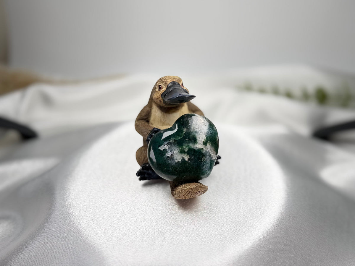 Treasured Energies small heart-shaped Ocean Jasper crystal held by a cute platypus figurine on a white satin surface.