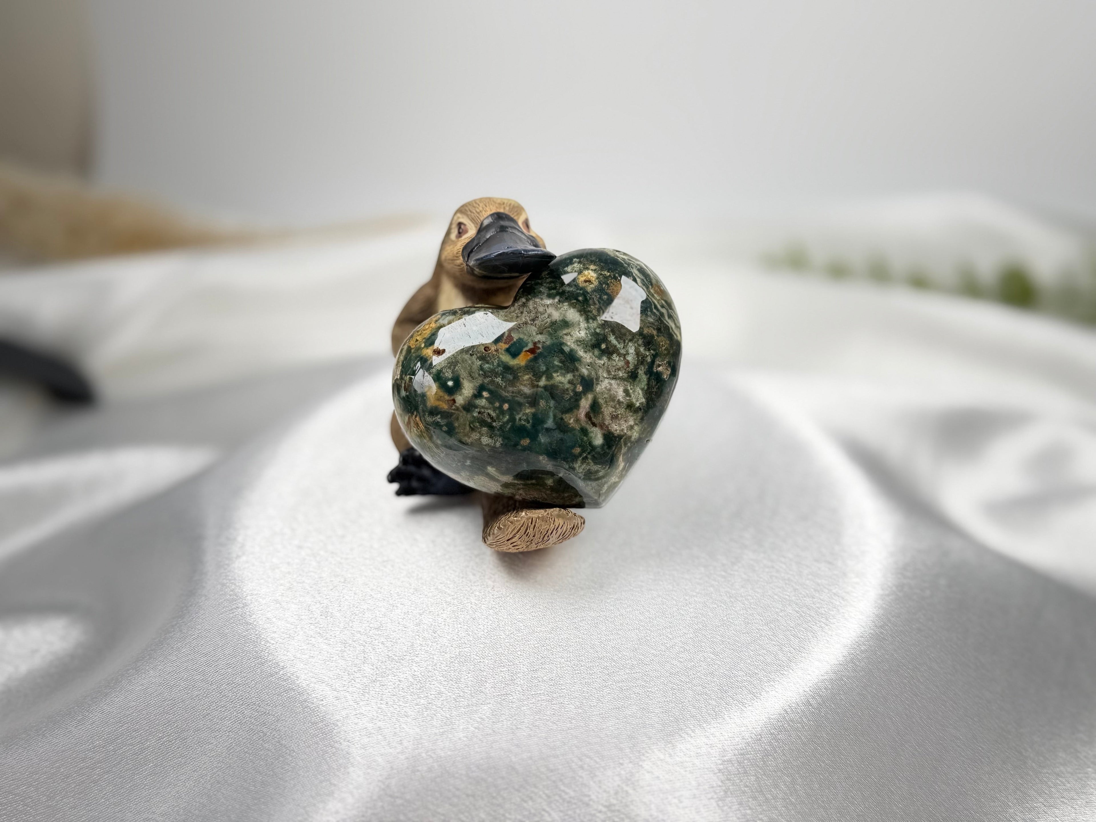 Treasured Energies large polished Ocean Jasper crystal heart held by a small platypus figurine on white satin fabric.