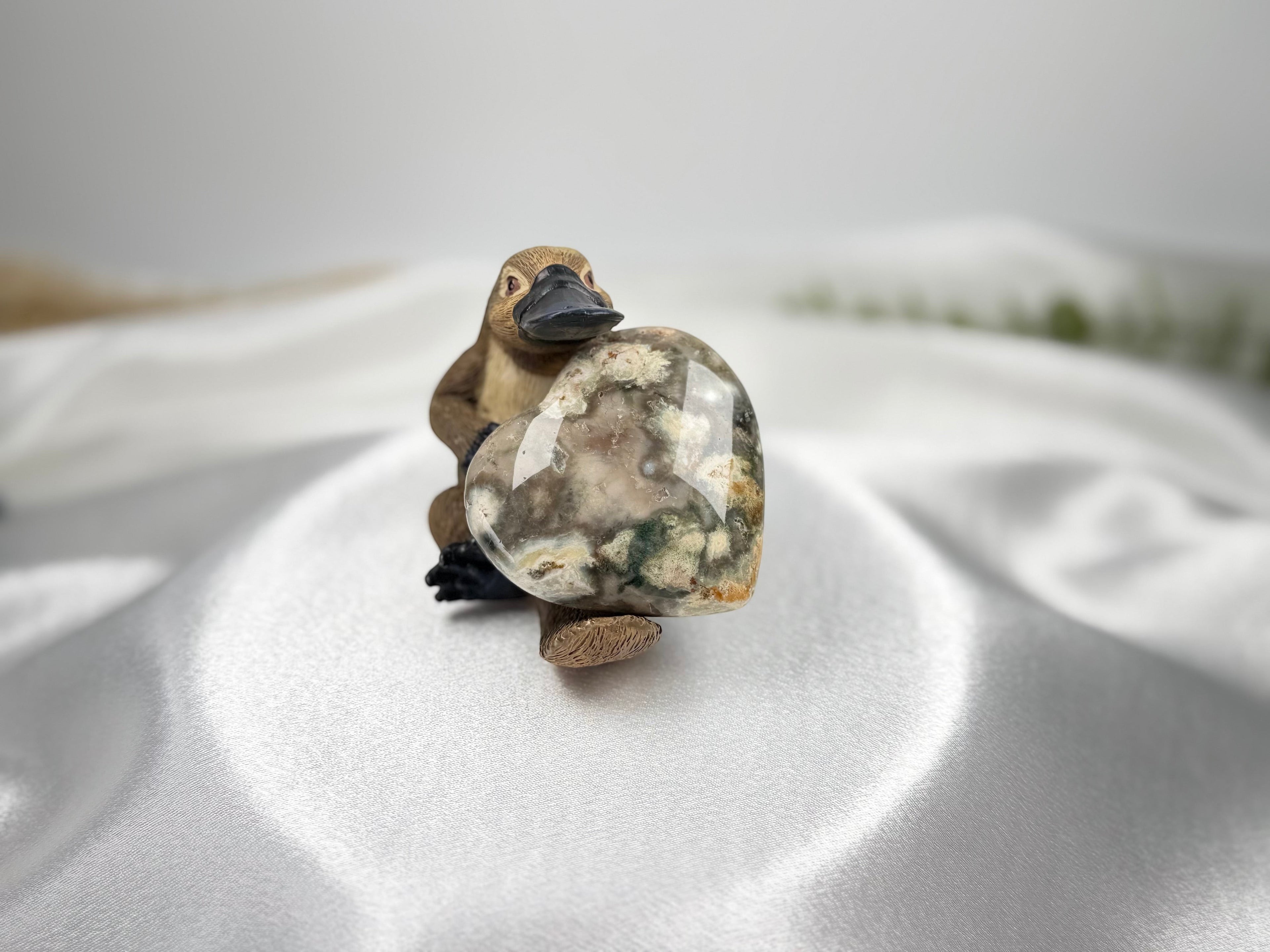 Treasured Energies large polished Ocean Jasper crystal heart held by a small platypus figurine on white satin fabric.