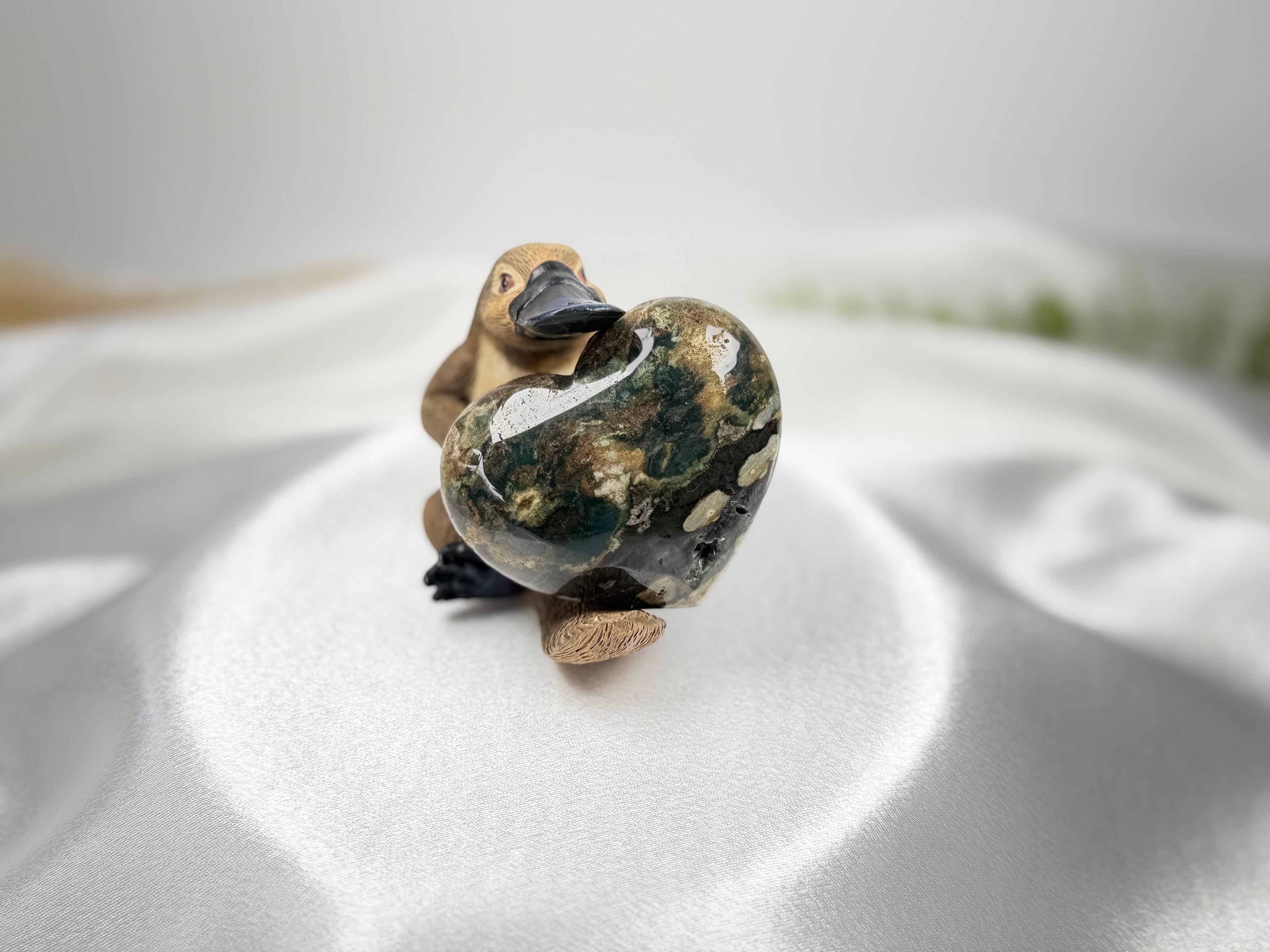 Treasured Energies large polished Ocean Jasper crystal heart held by a small platypus figurine on white satin fabric.