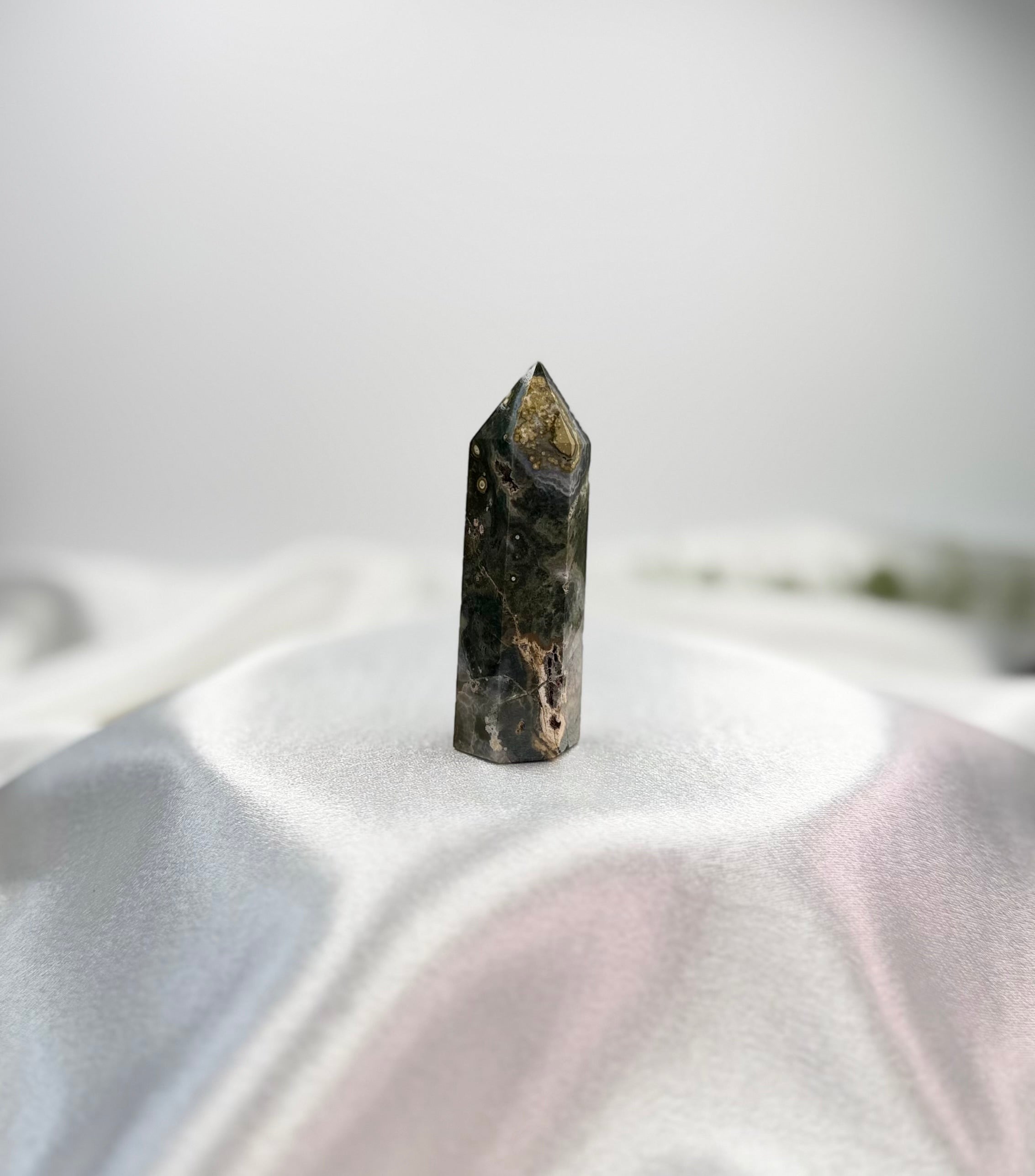 Treasured Energies Ocean Jasper crystal tower with dark green and brown patterns on a white satin fabric background.