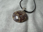 Treasured Energies polished orbicular poppy jasper pendant with brown and tan spots on a black cord against a textured cloth.