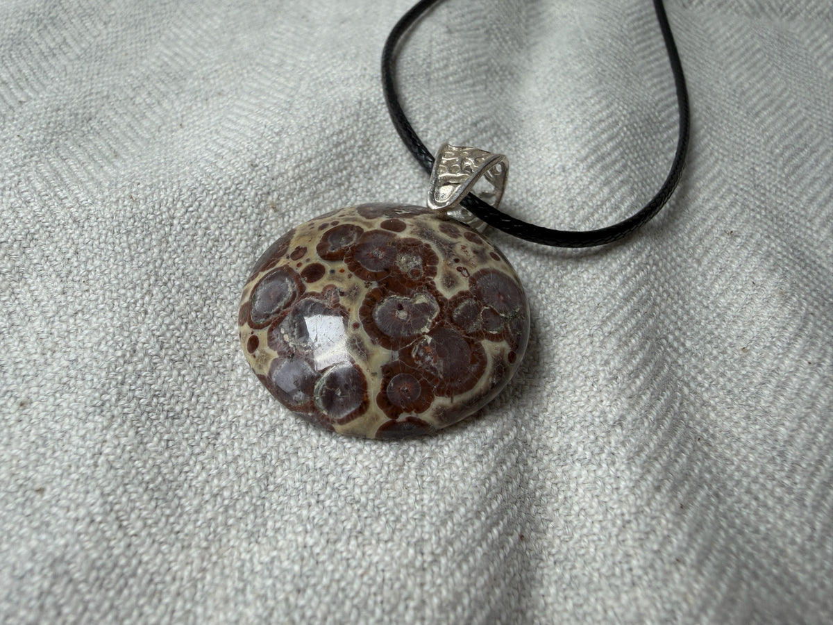 Treasured Energies polished orbicular poppy jasper pendant with brown and tan spots on a black cord against a textured cloth.