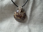 Treasured Energies round orbicular poppy jasper pendant with chocolate and tan spots on a black cord against linen fabric