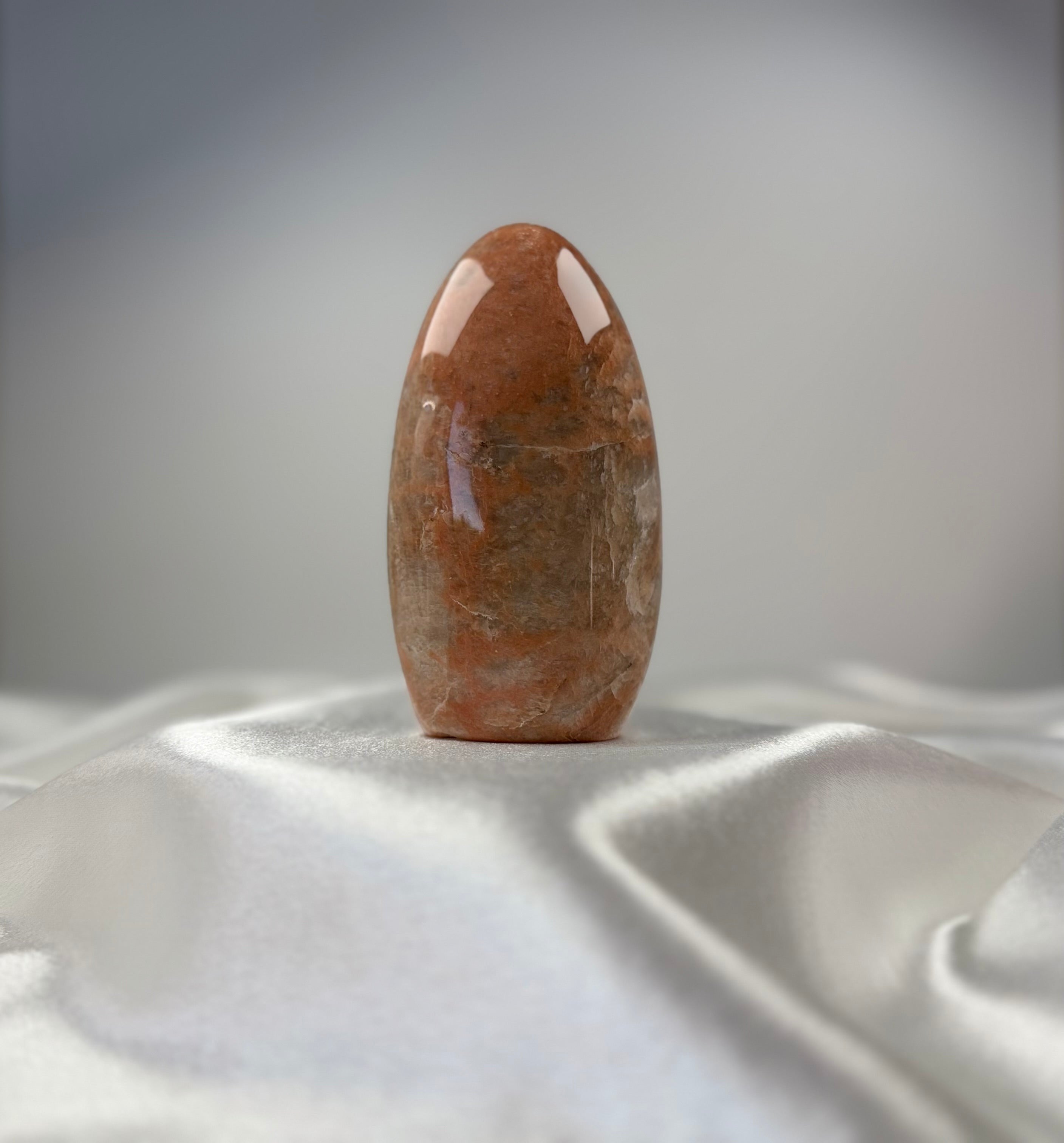 Treasured Energies polished peach moonstone crystal freeform with smooth, organic surface on a white satin fabric.