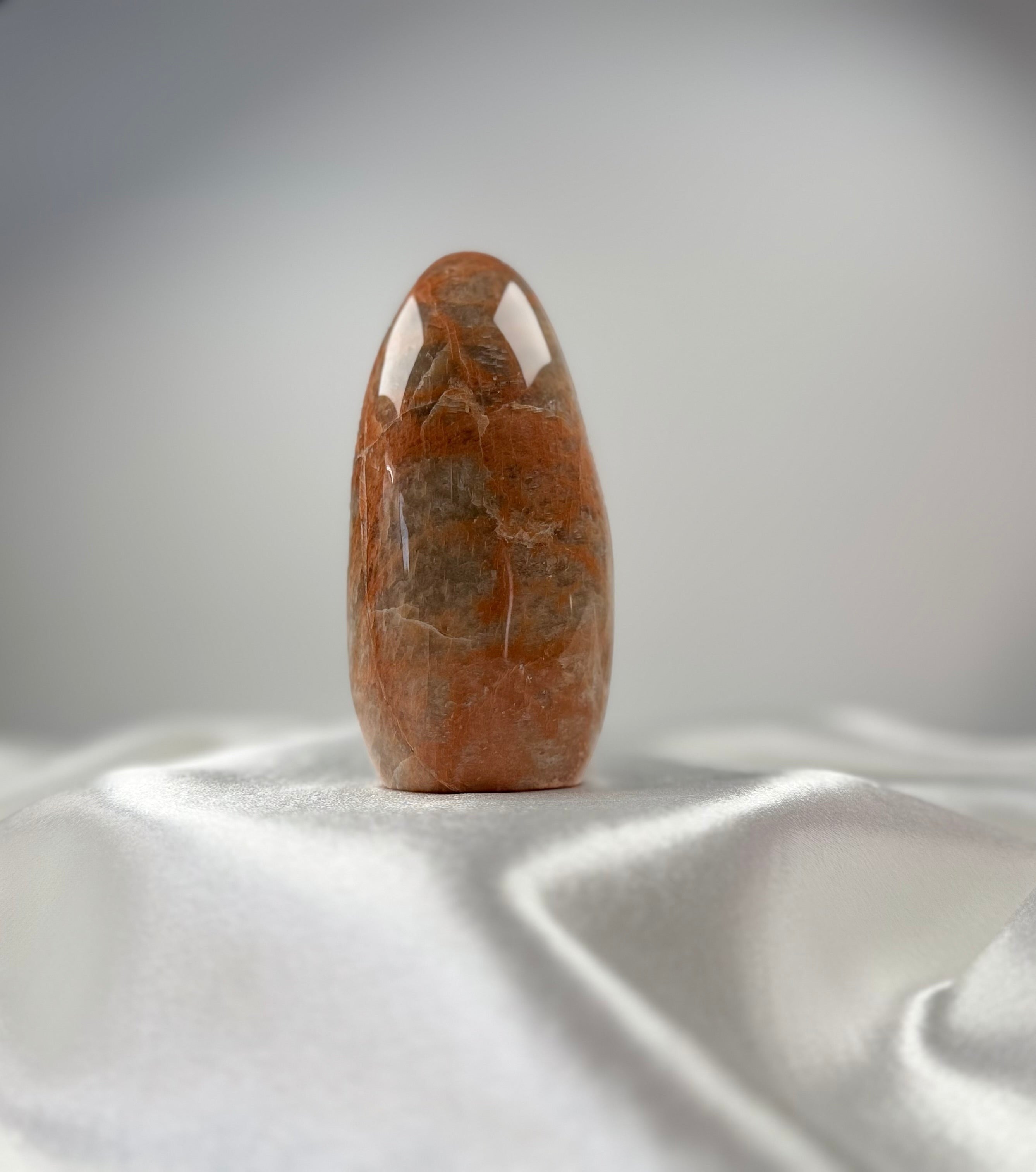 Treasured Energies polished peach moonstone crystal freeform with organic banding on a white satin background.