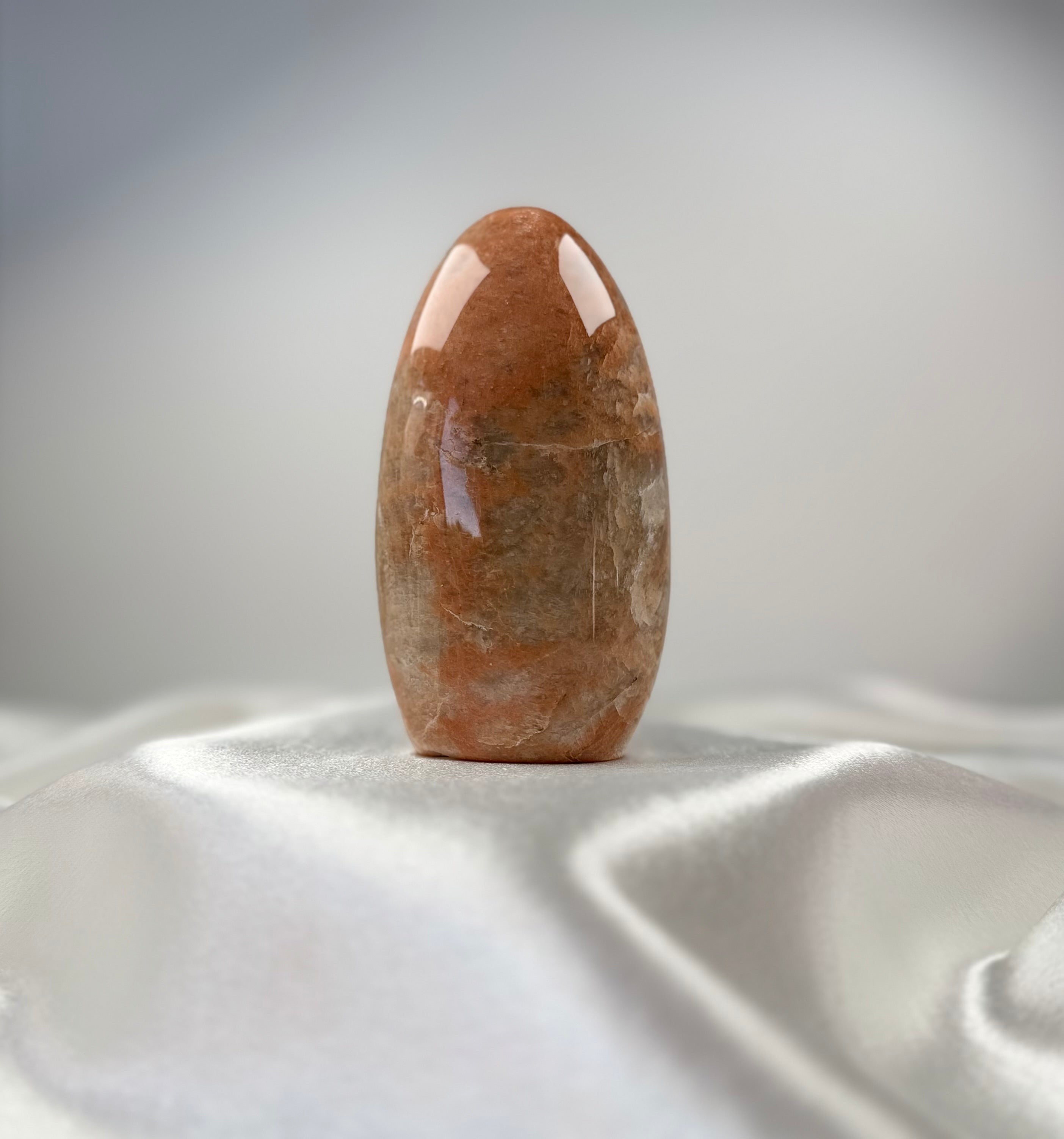 Treasured Energies polished peach moonstone crystal freeform with organic banding on a white satin fabric background.