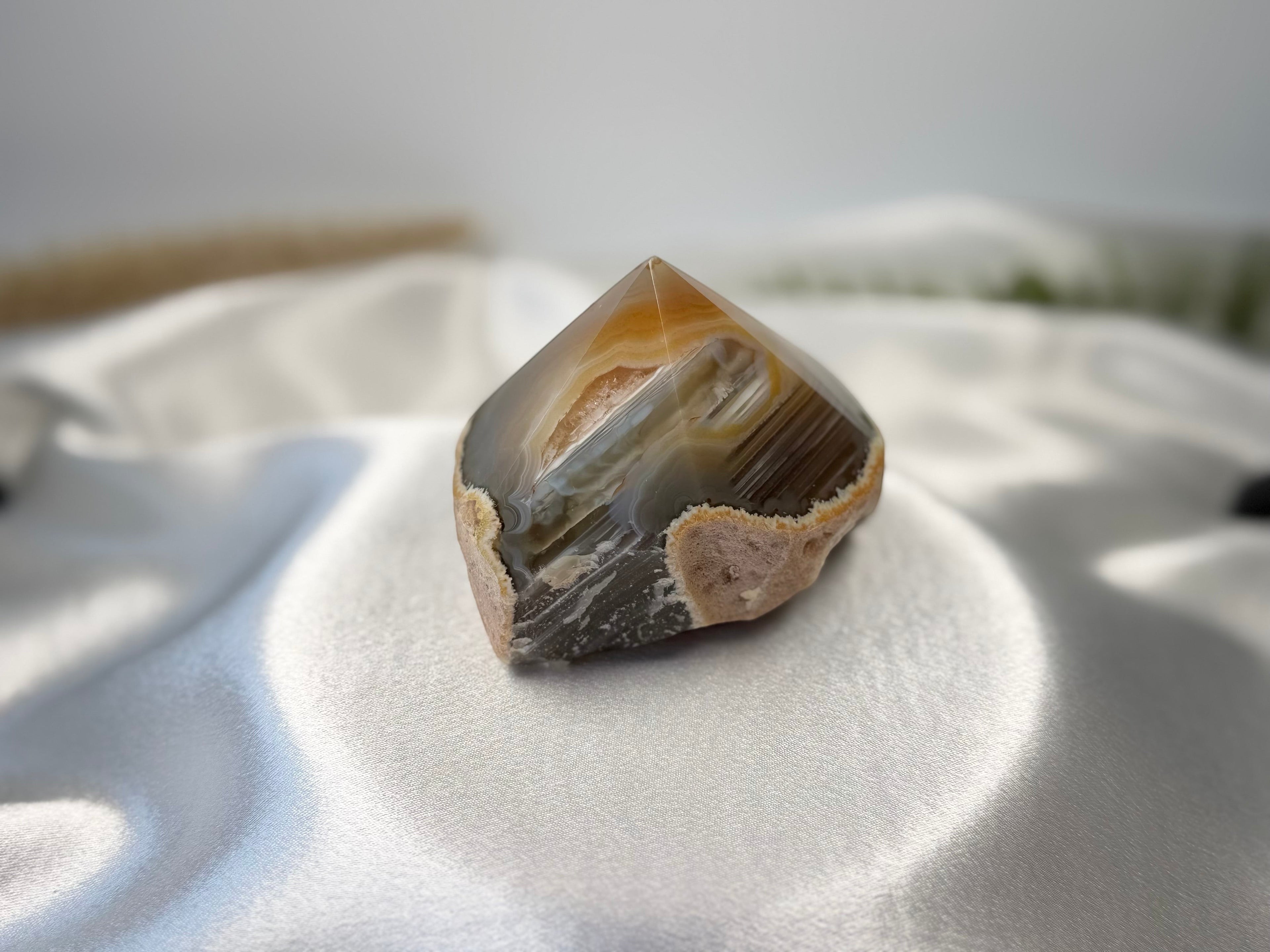 Treasured Energies polished agate crystal point with natural brown and grey banding on a white satin background.