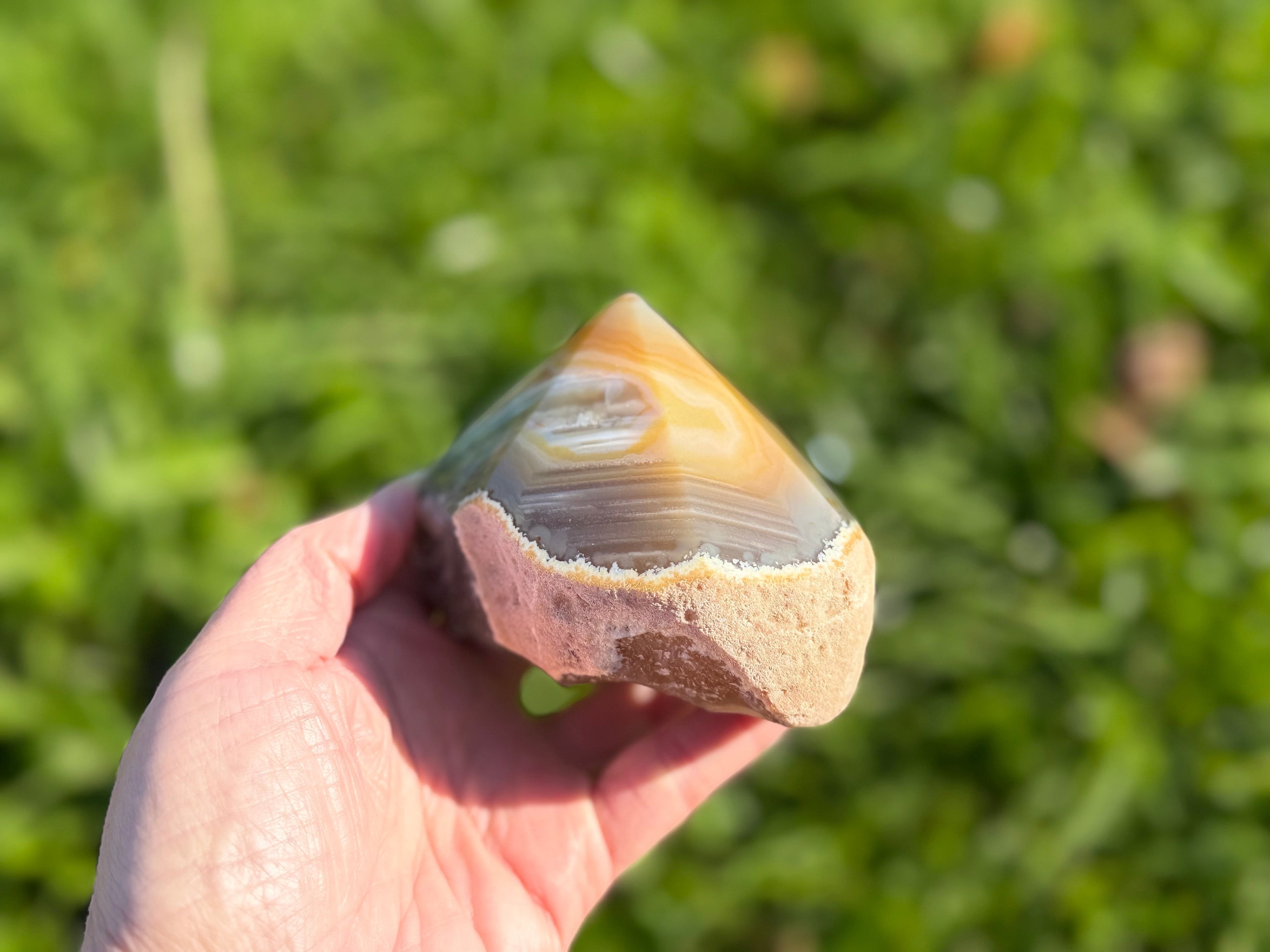 Treasured Energies polished agate point with natural brown and grey banding held in a hand against a green background.
