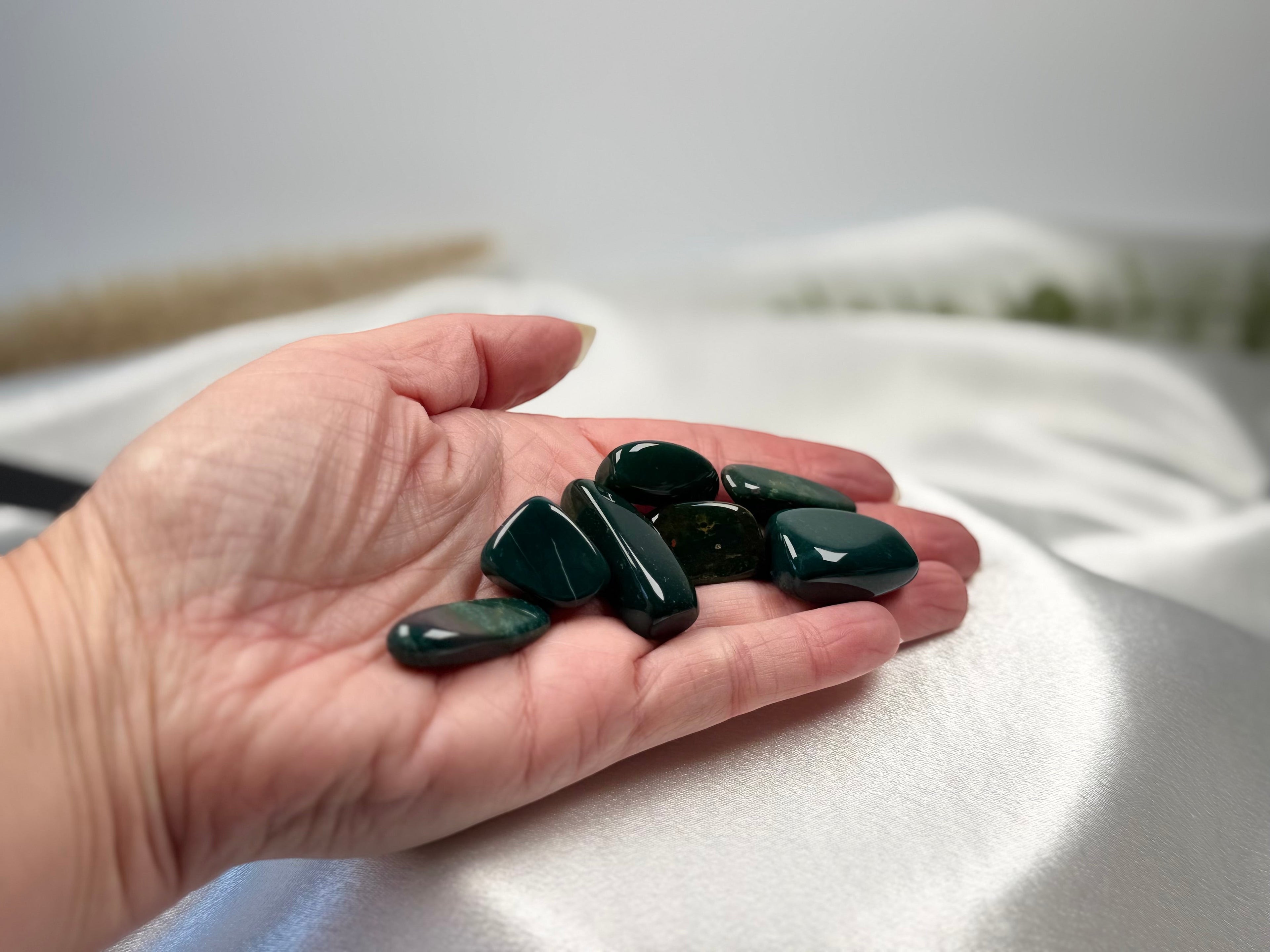 Treasured Energies polished bloodstone tumbled gemstones held in a palm against a soft white silk background.