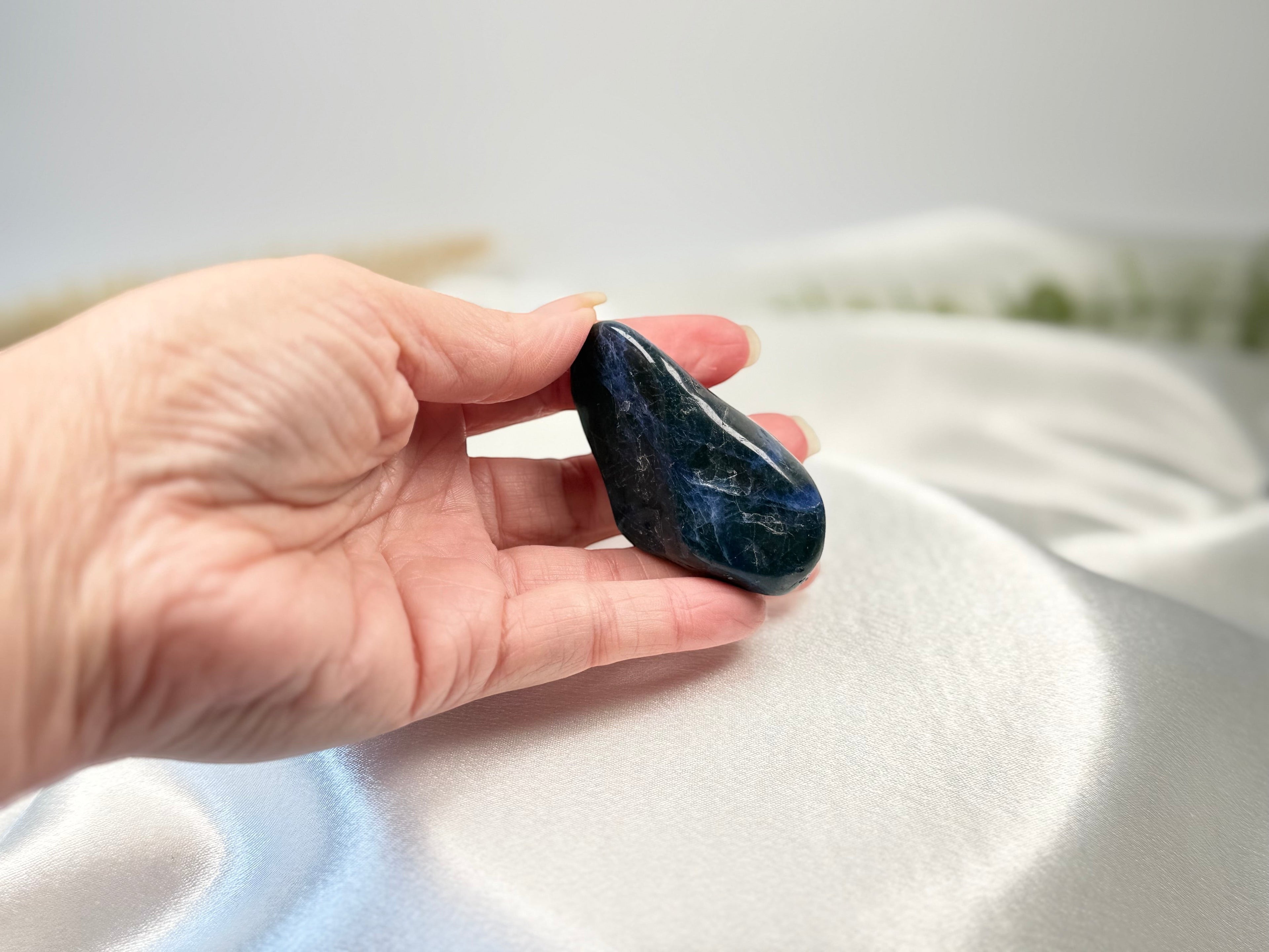 Treasured Energies polished royal blue sodalite tumbled stone held in a hand against a soft white satin background.