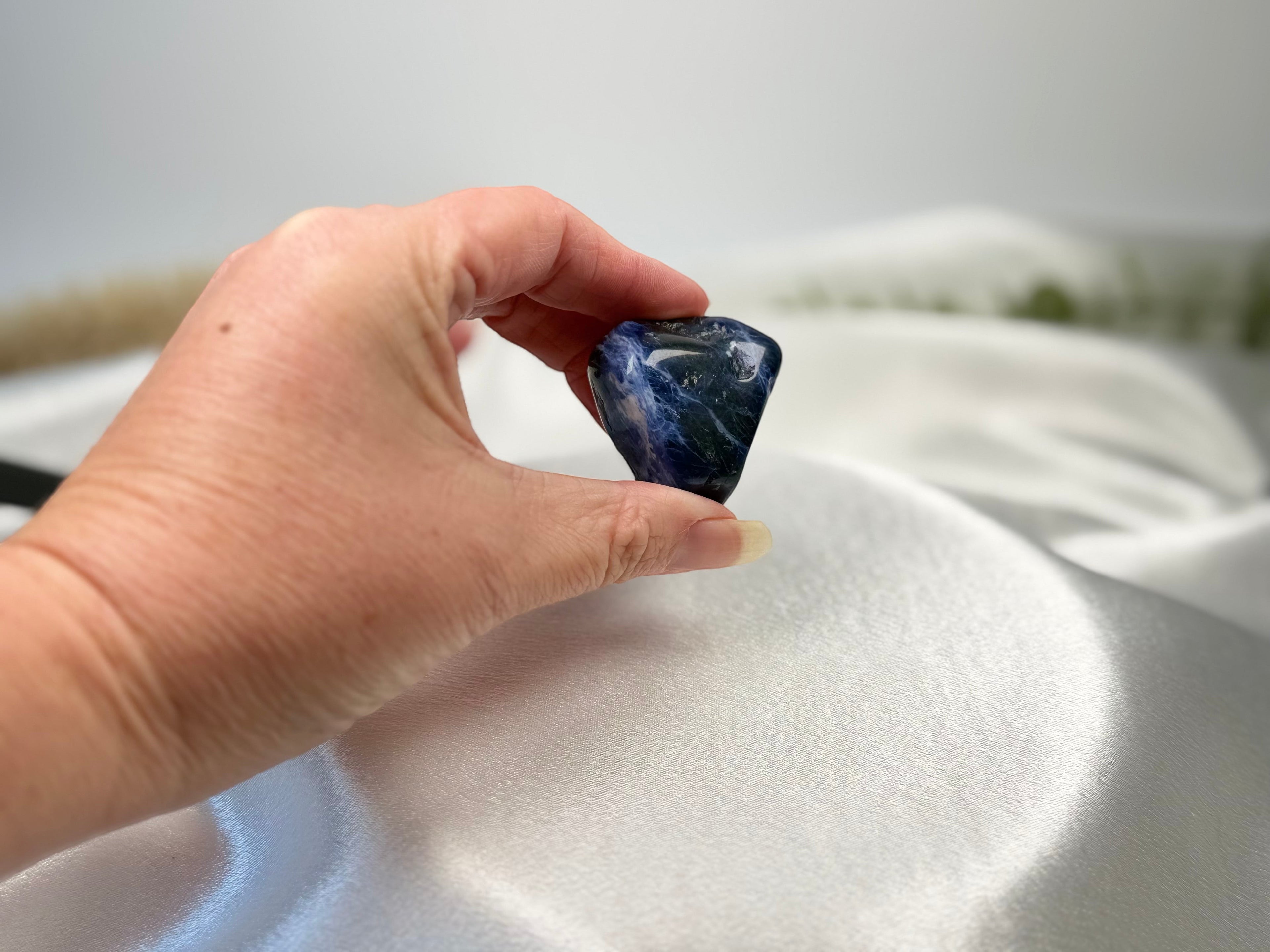 Treasured Energies polished royal blue sodalite tumbled stone held in a hand against a white satin background.