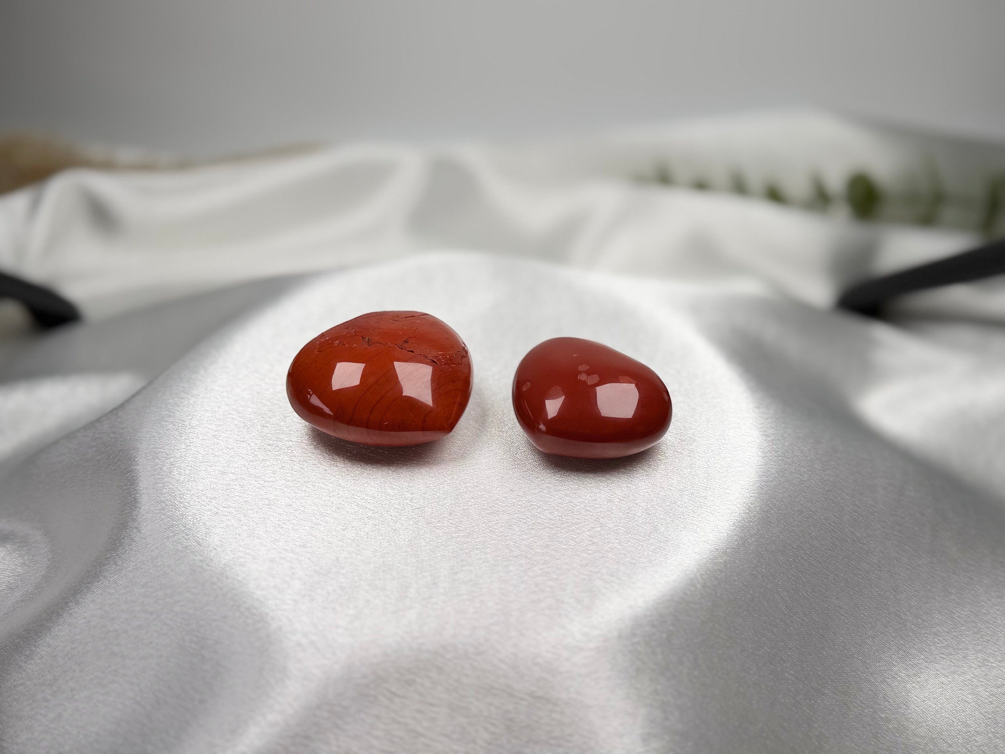 Two polished heart-shaped Red Jasper crystals from Treasured Energies resting on white satin fabric.