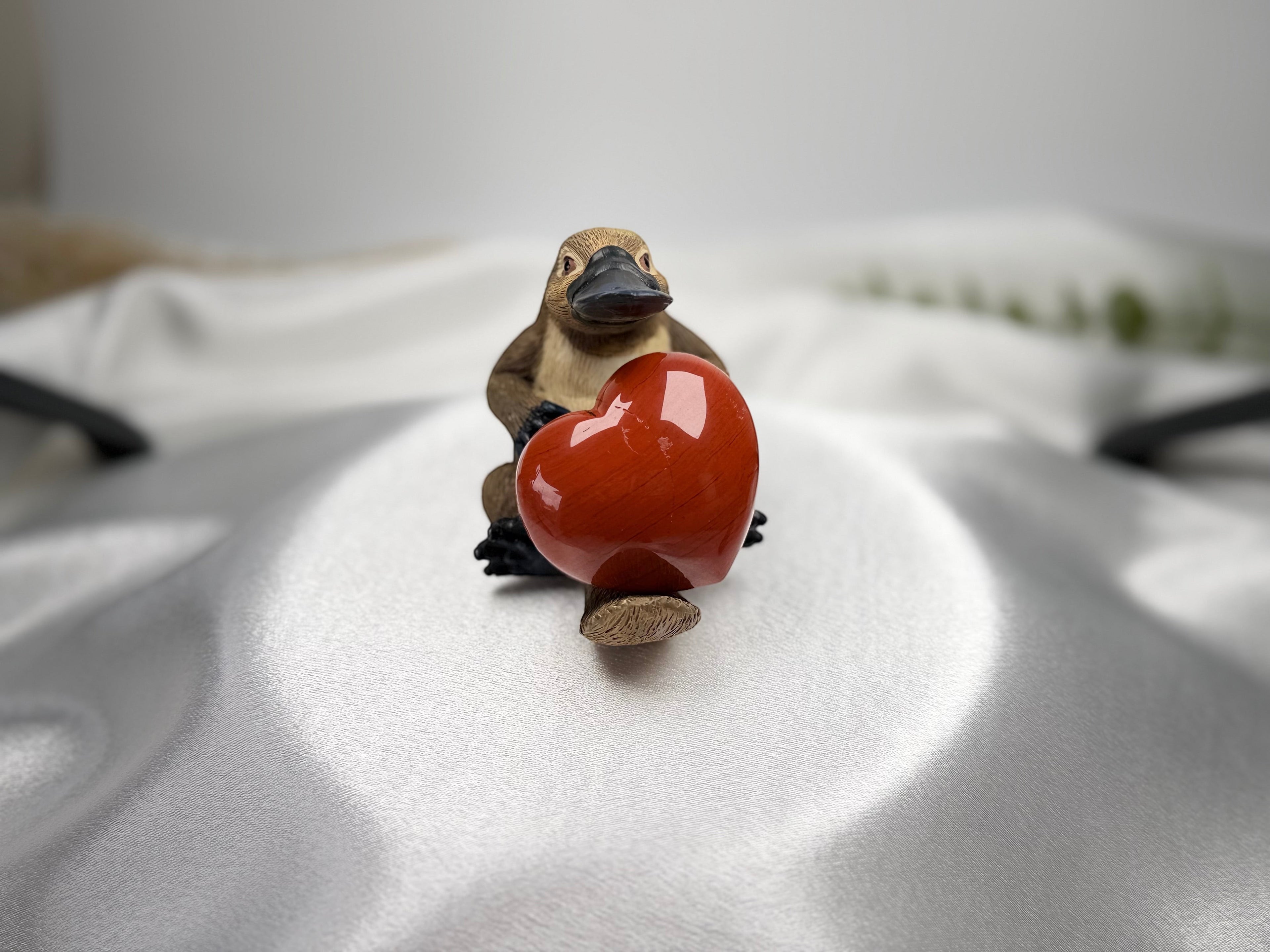 Treasured Energies red jasper heart crystal held by a small platypus figurine on a white satin background.