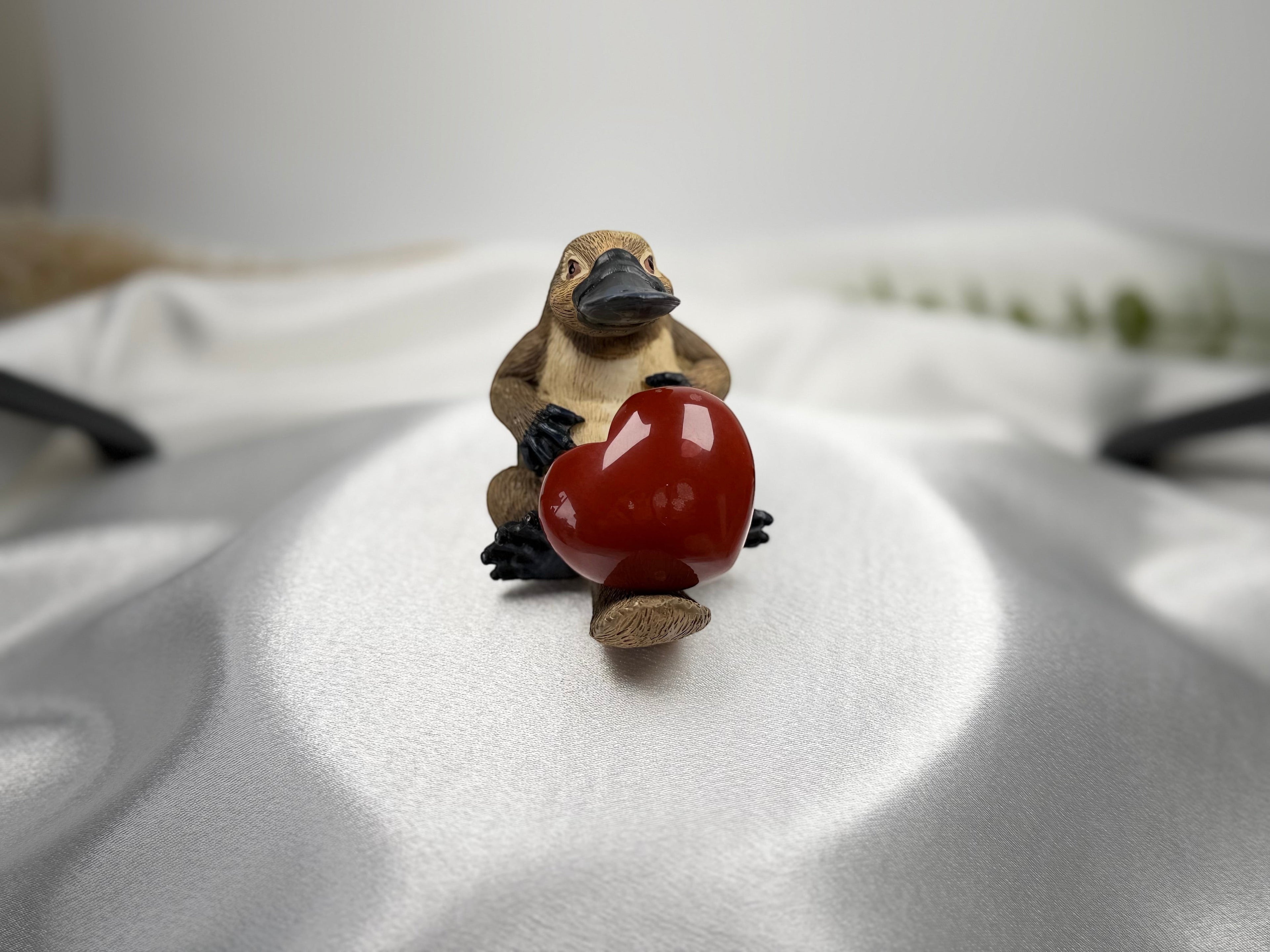 Treasured Energies red jasper crystal heart held by a small platypus figurine on a white satin background.