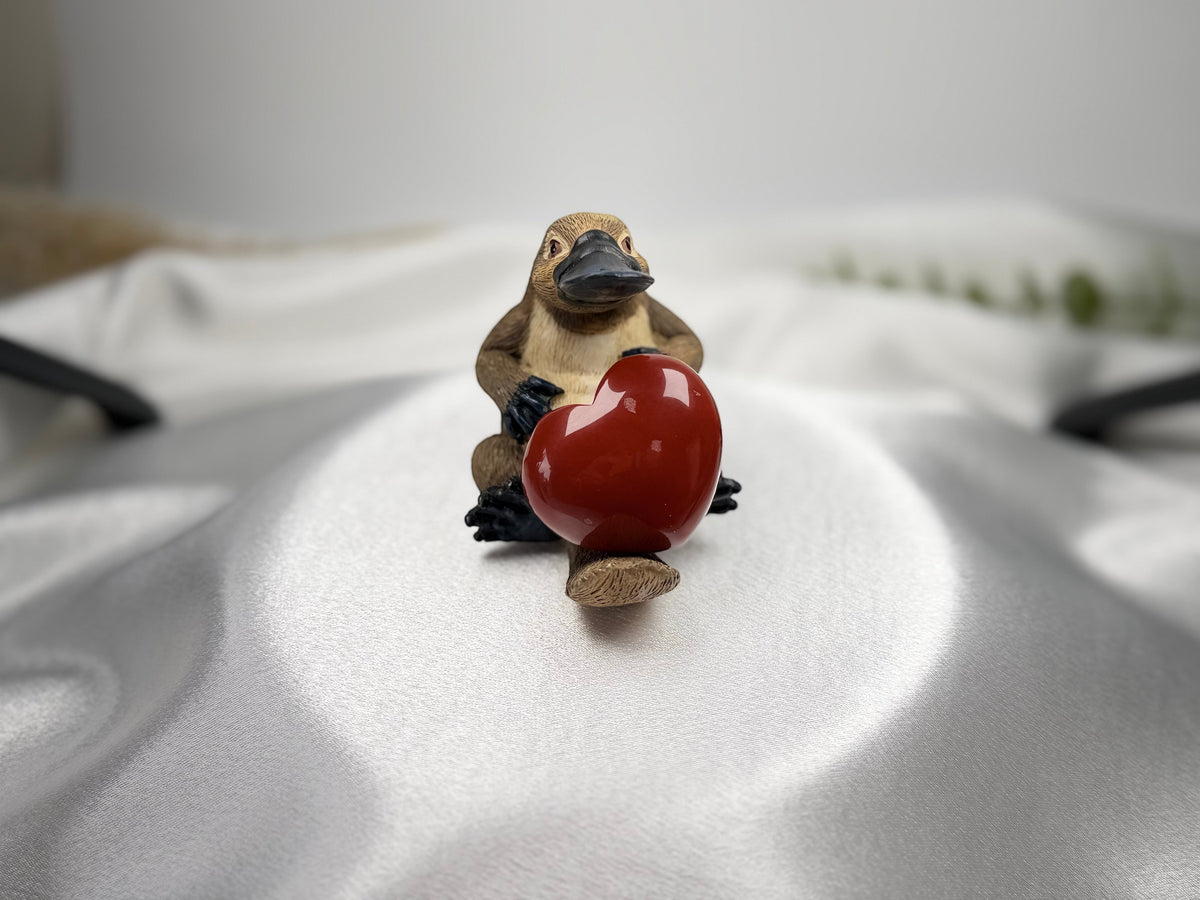 Treasured Energies red jasper heart held by a small platypus figurine on a smooth white satin background