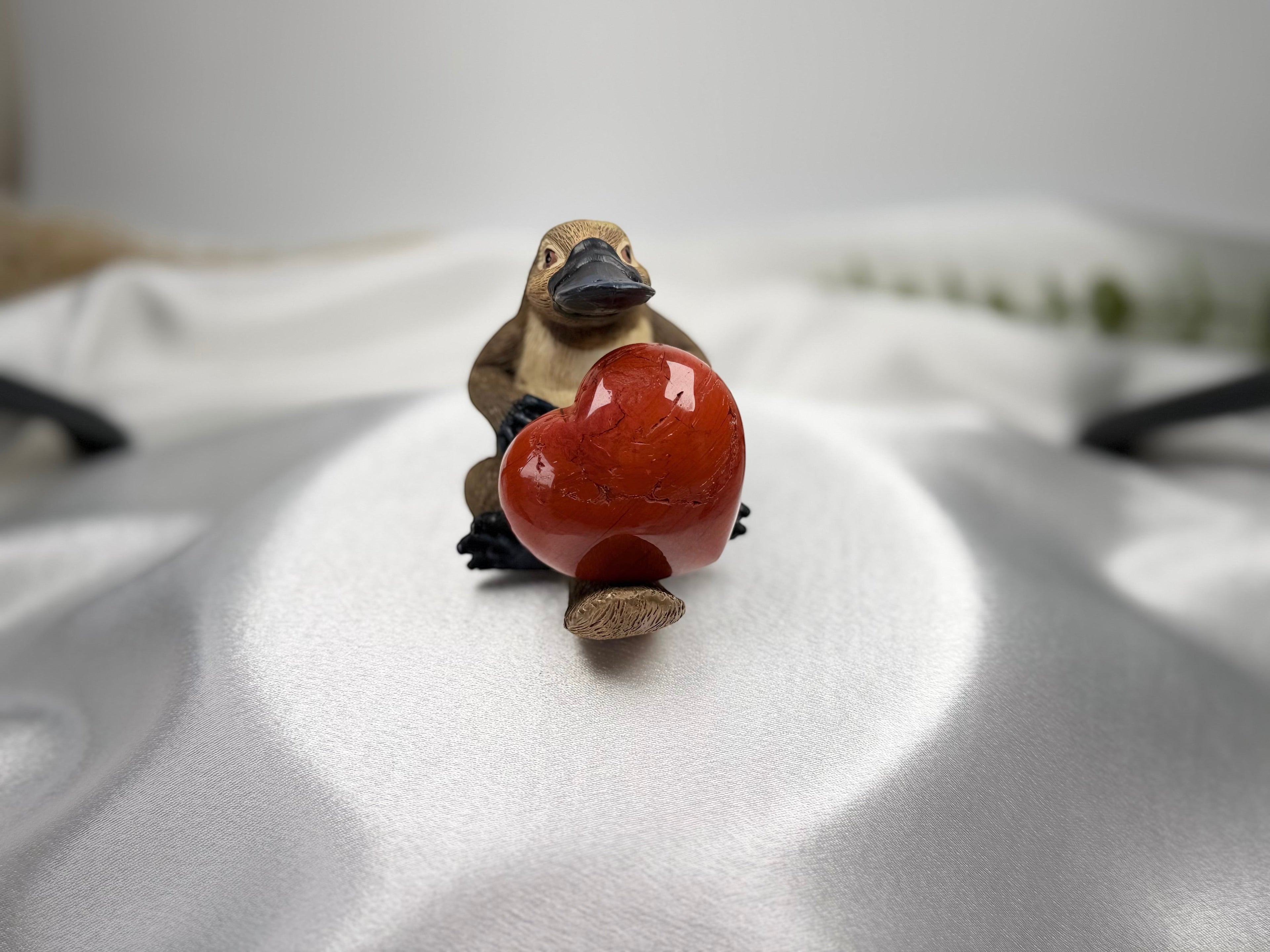 Treasured Energies polished red jasper crystal heart held by a small platypus figurine on a white satin background.