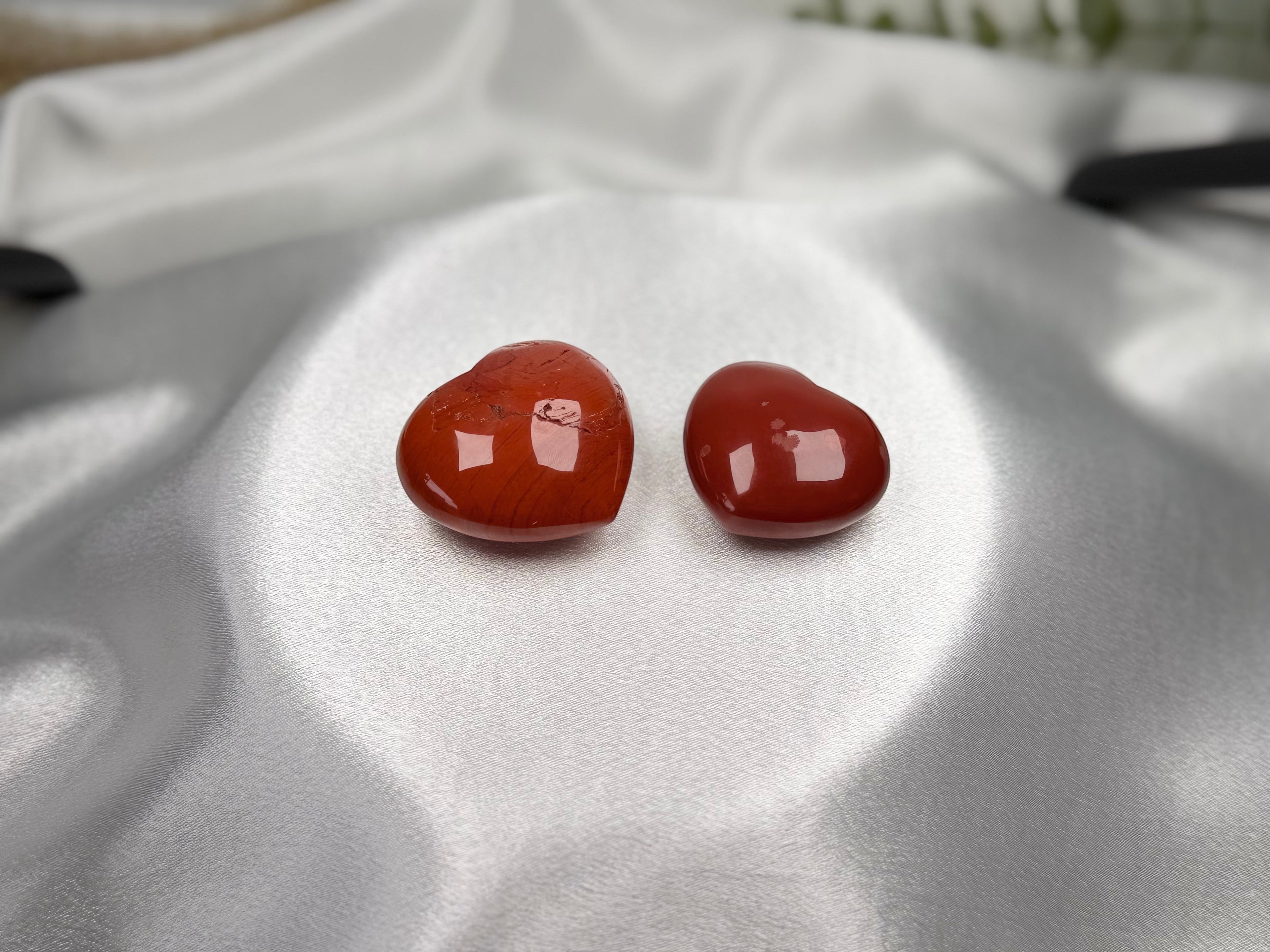 Two polished red jasper heart-shaped crystals displayed on white satin fabric by Treasured Energies.