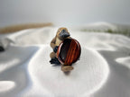 Treasured Energies red tiger's eye worry stone held by a small platypus figurine on a white satin background.