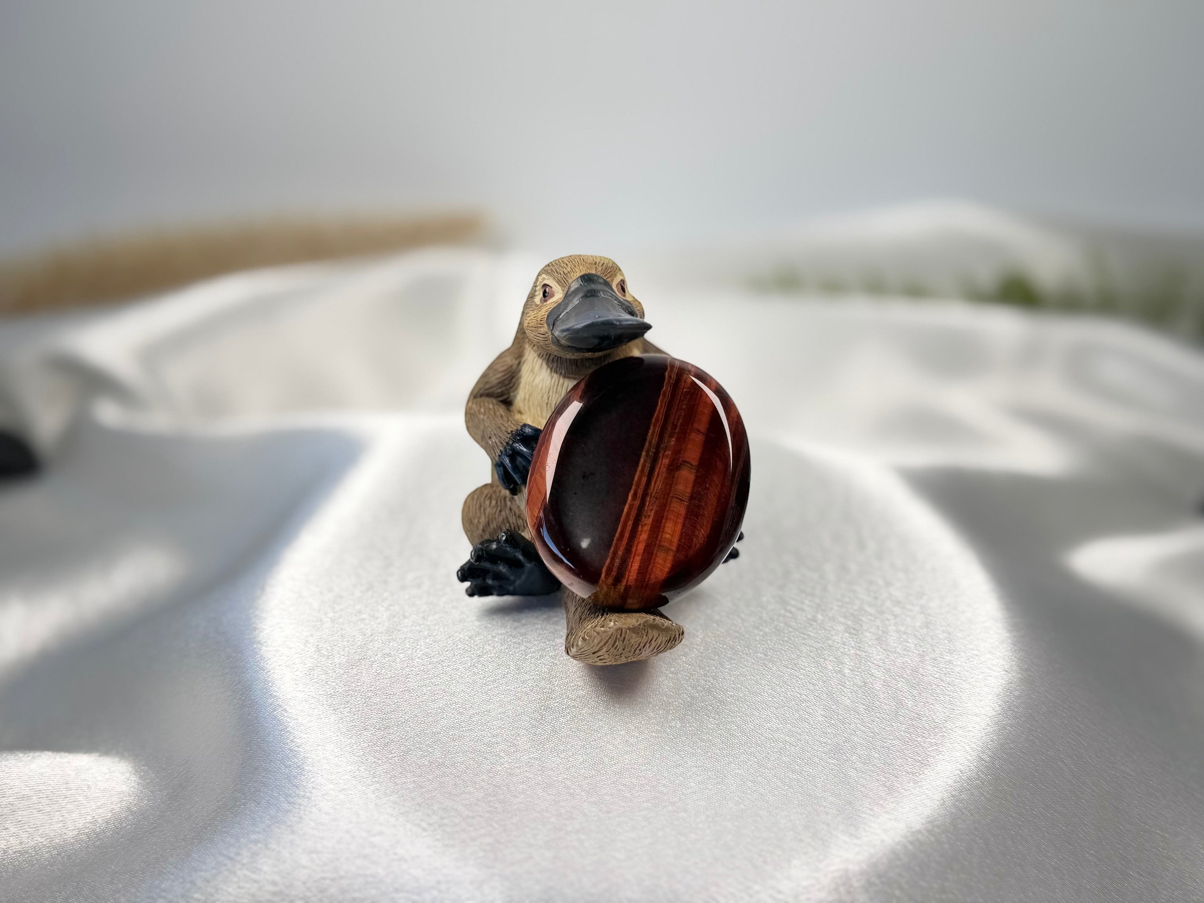 Treasured Energies red tiger's eye worry stone held by a small platypus figurine on a white satin background.