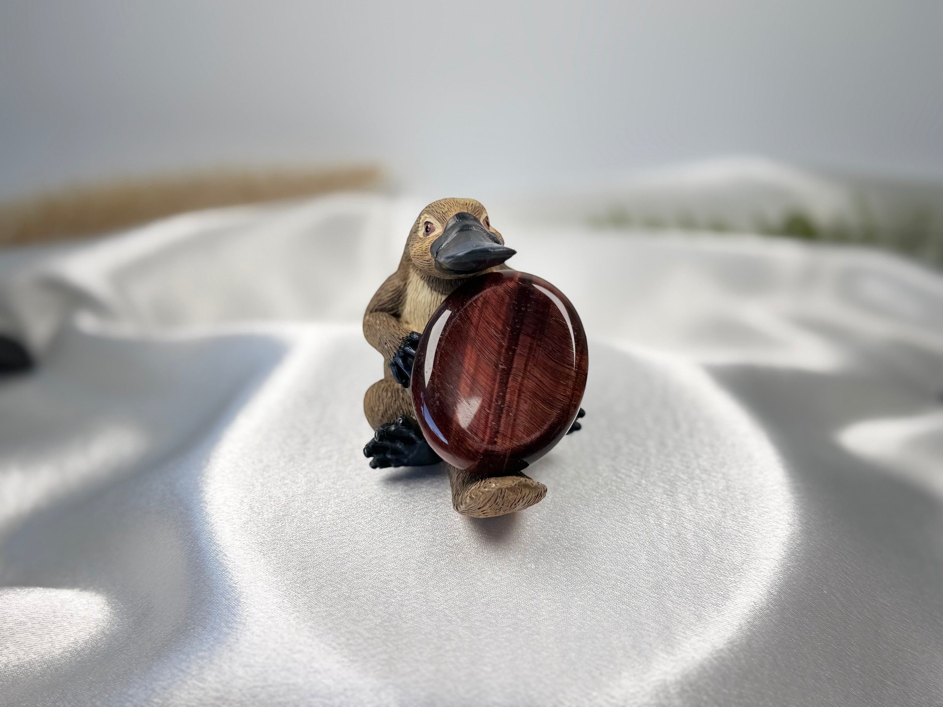 Treasured Energies red tiger's eye worry stone held by a small platypus figurine on a white satin fabric background.
